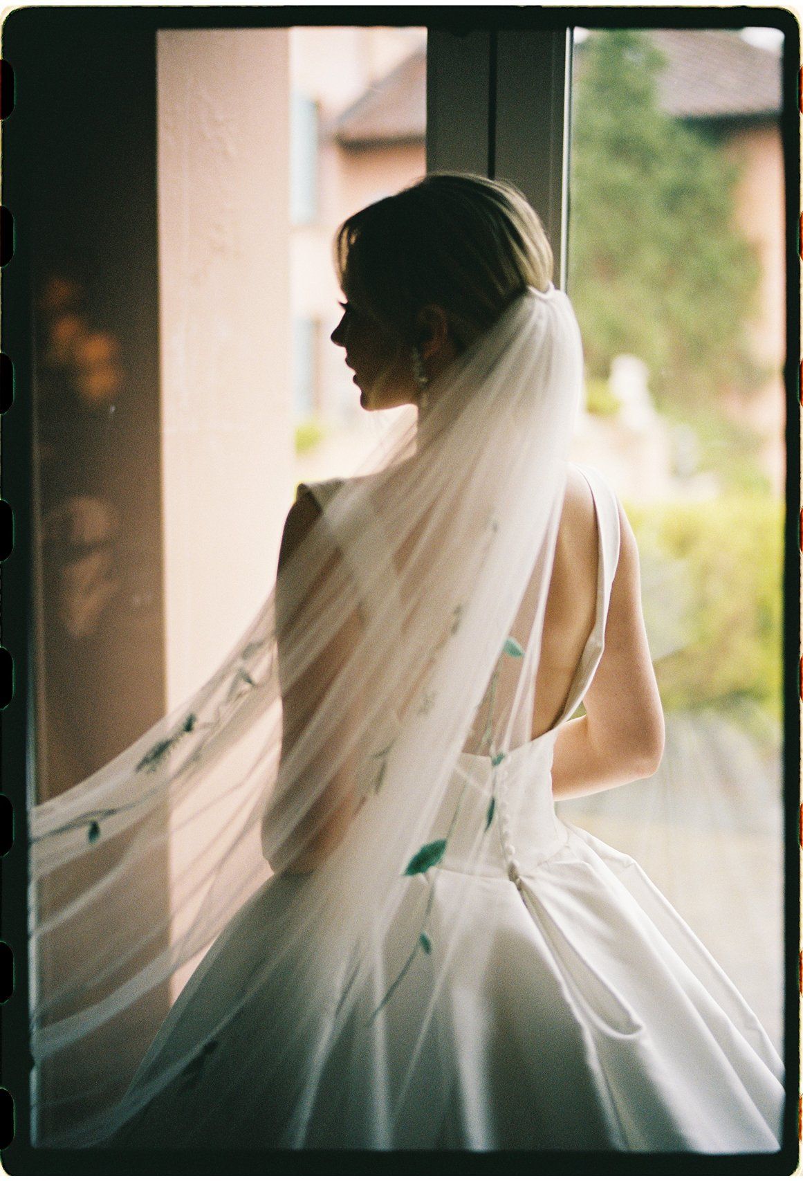 Bride in white gown and flowing veil stands by sunlit window, showcasing elegant backless dress with green floral details.