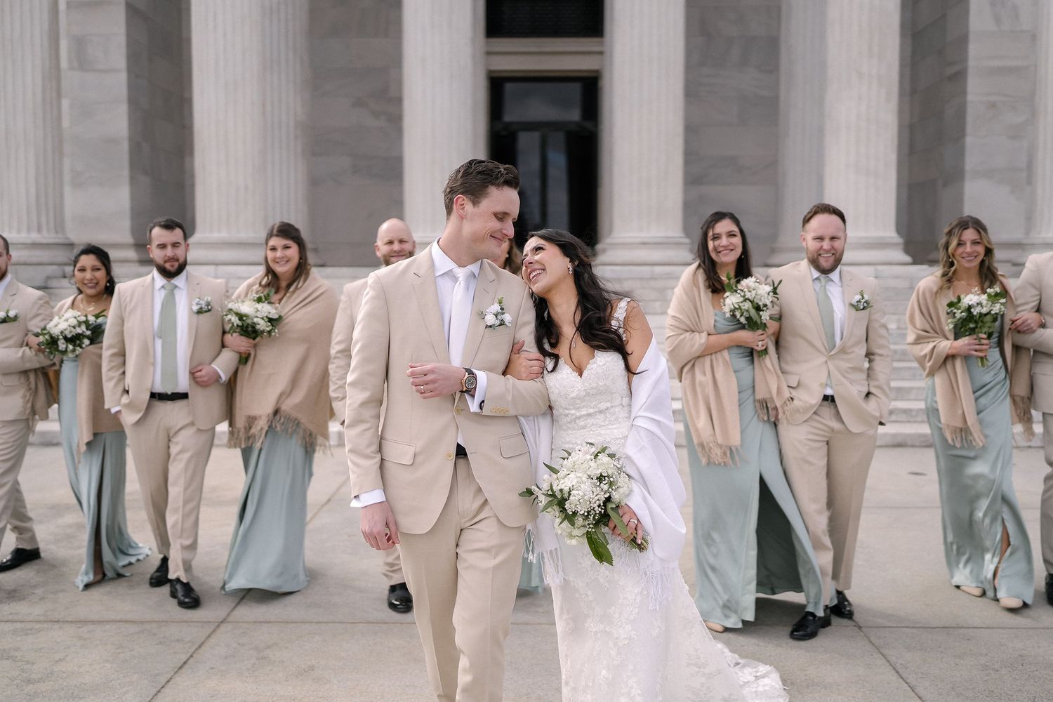 Bride and groom walk together in front of columns with wedding party in sage and tan attire holding white bouquets.