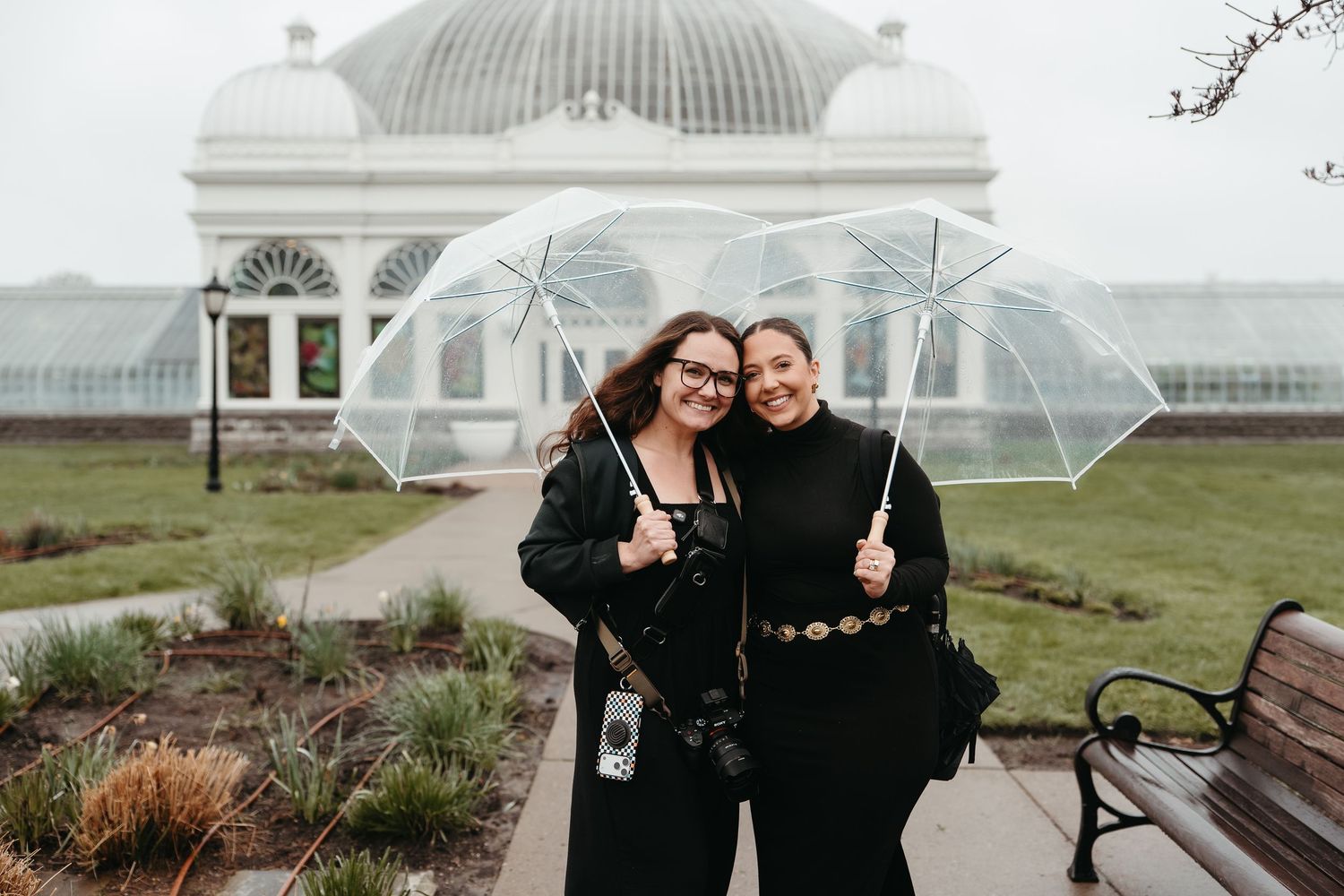 Two women holding clear umbrellas smiling outside a glass greenhouse on cloudy day.