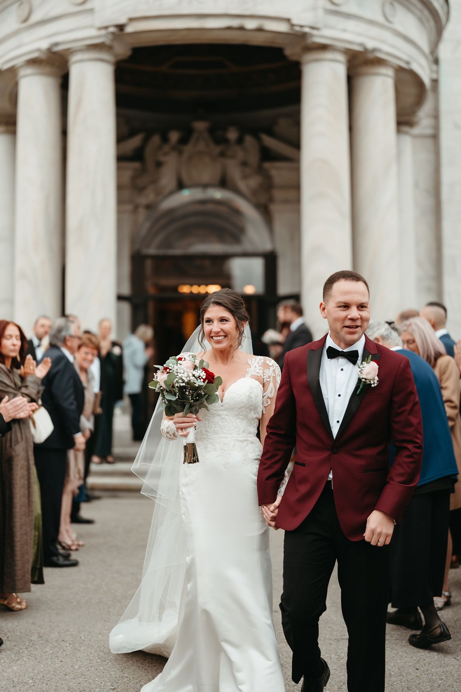 Newlywed couple walks down aisle after wedding ceremony, bride in white gown with bouquet, groom in burgundy tuxedo jacket.