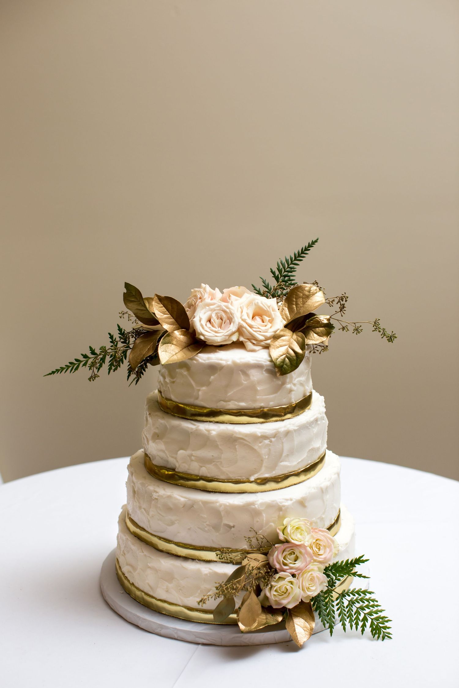 A four-layer wedding cake with butter cream frosting and gold ribbon around it, decorated with beige roses and gold leaves.