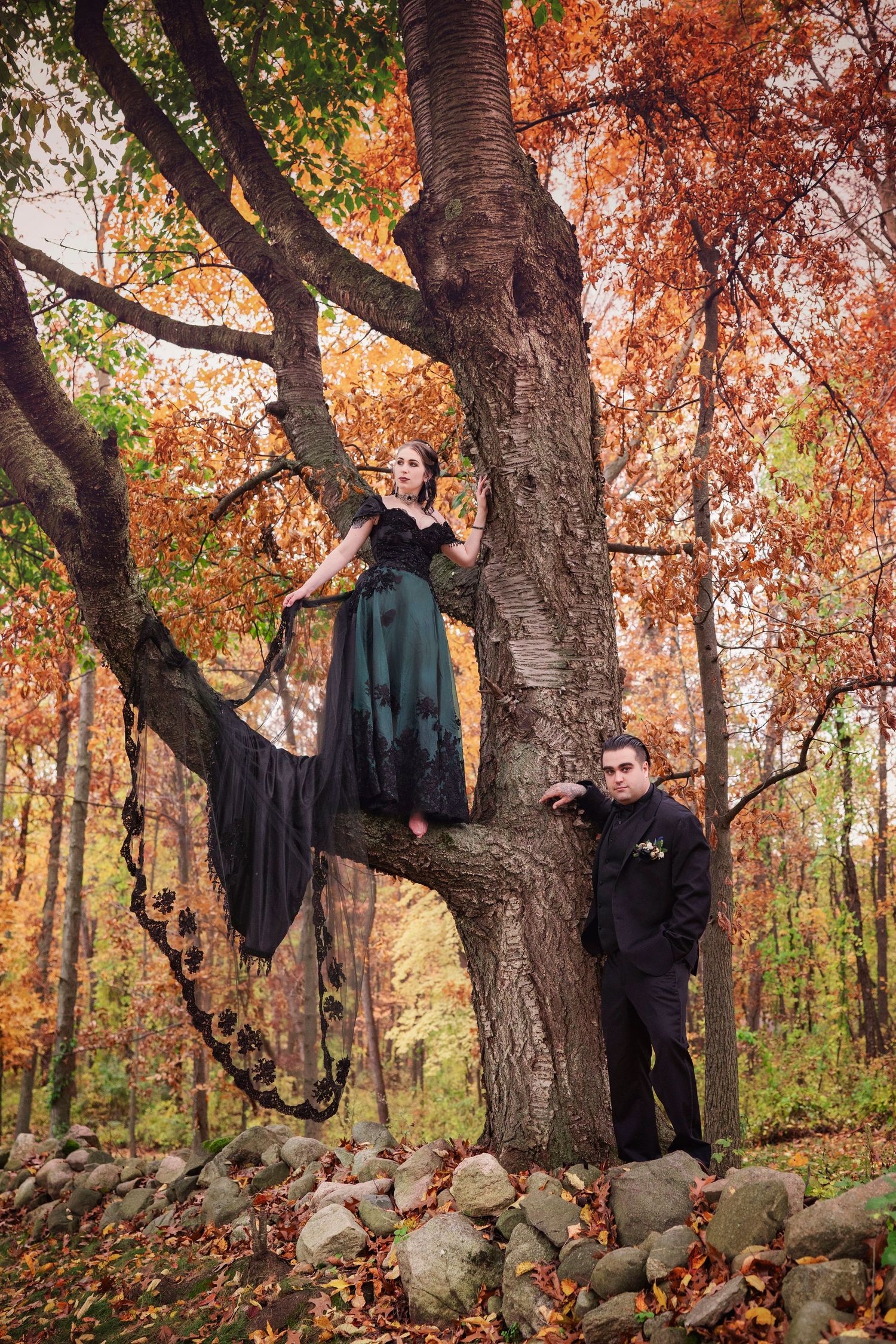 A couple poses for their Halloween-themed wedding in a Michigan forest. The bride stands in a tree with a black gown, her train drapes low.