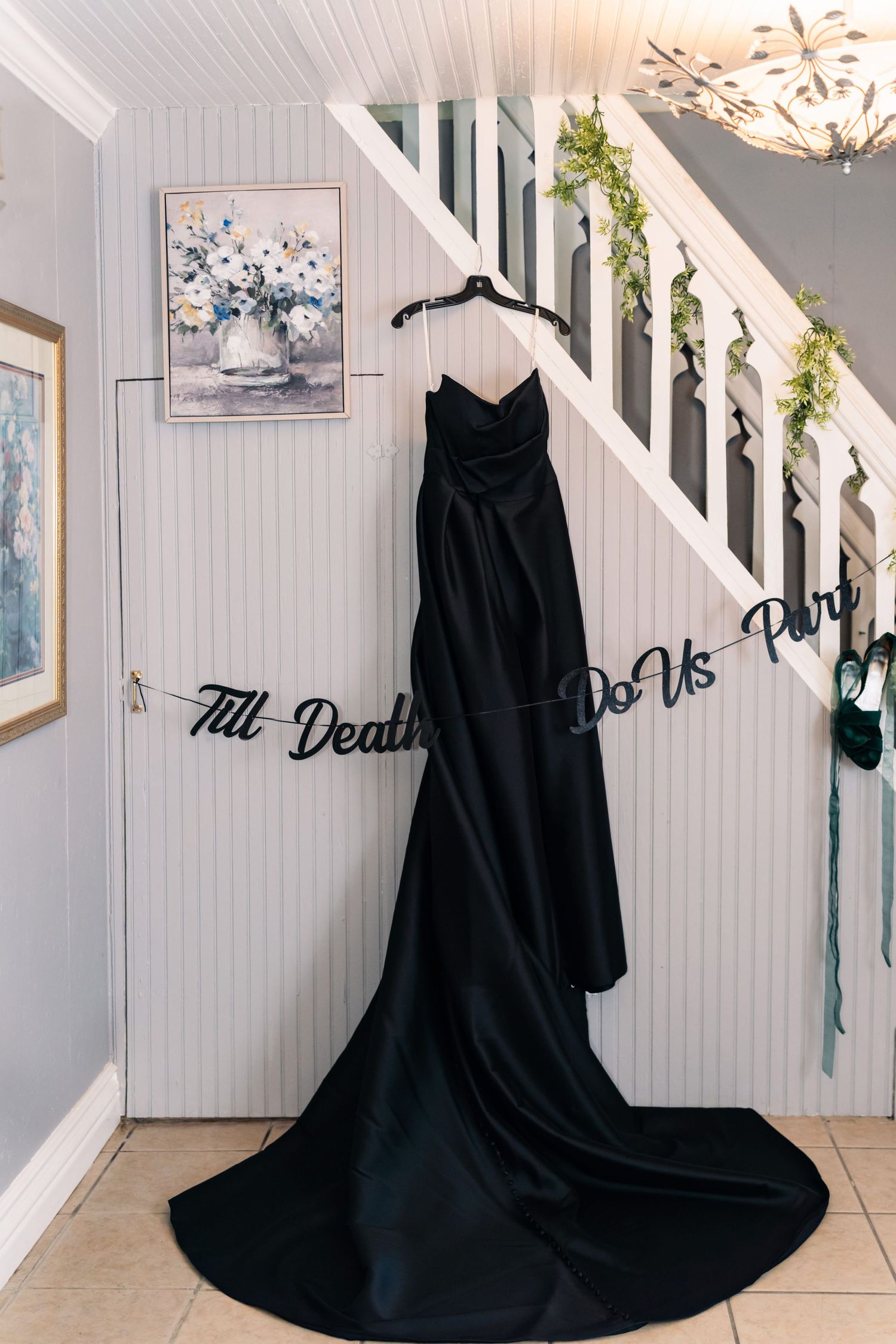 A black satin wedding dress with a long train hangs beneath a staircase, with a 'Till Death Do Us Part' sign adding a gothic-romantic touch.