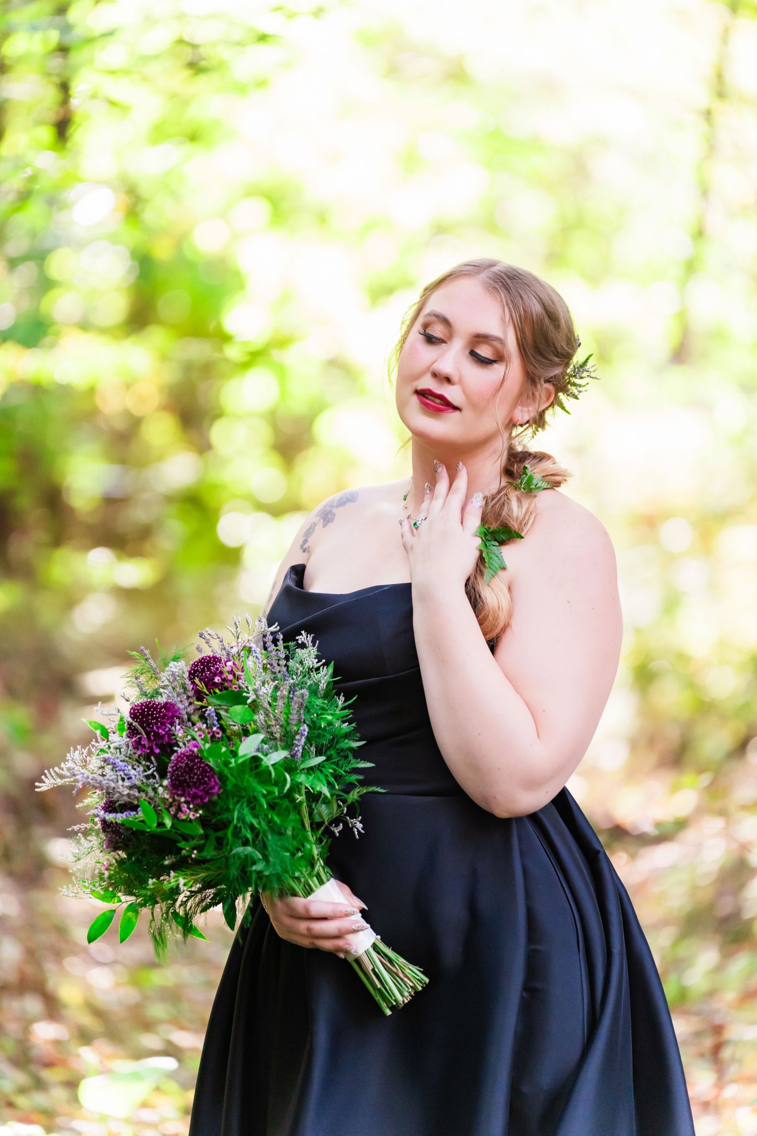 An auburn-haired bride in a black dress poses with a natural sun-drenched forest as a backdrop at Stonegate Manor in St Joseph Michigan.