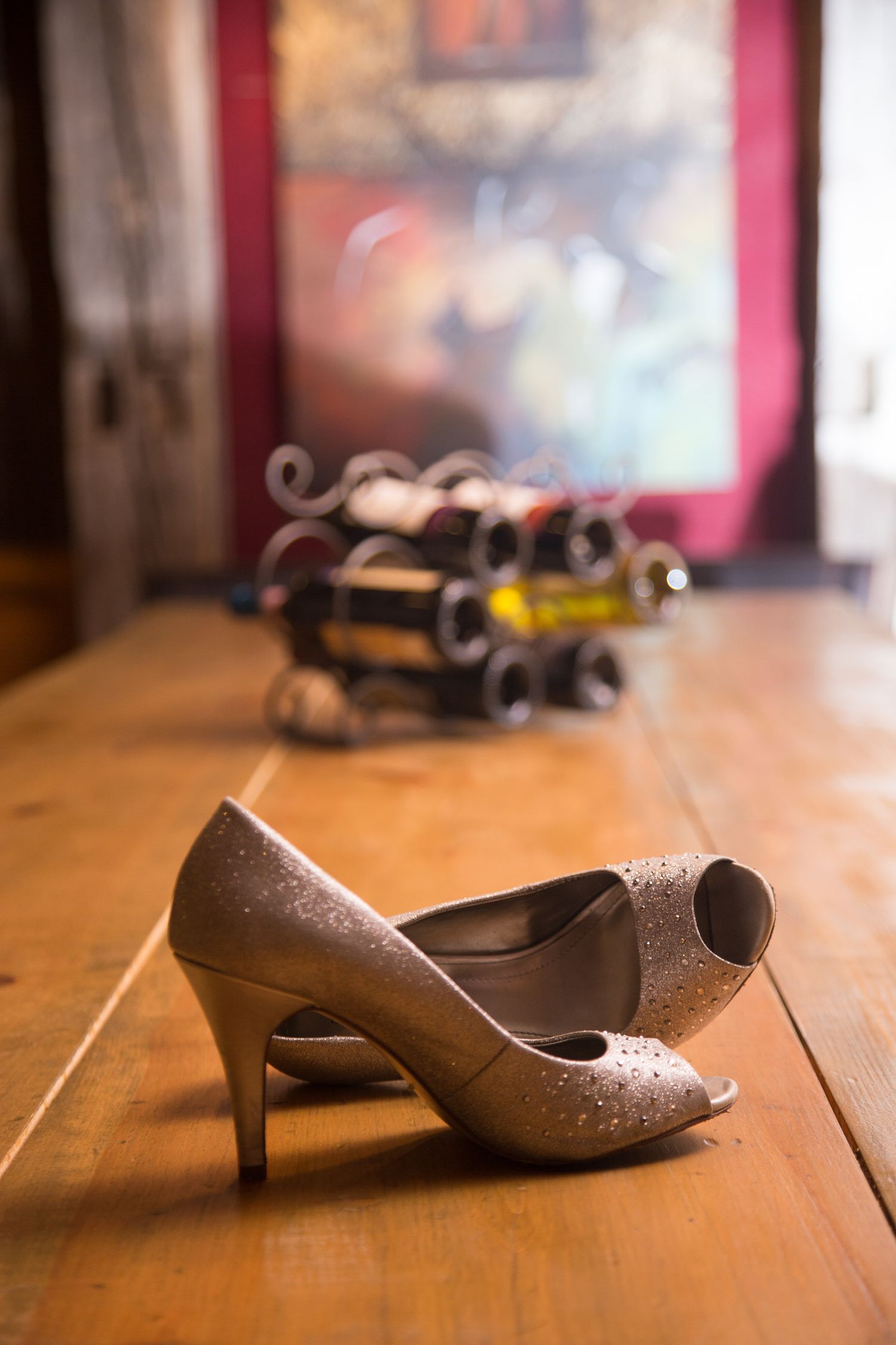Glittered taupe wedding day shoes lay casually in a wine cellar in front of a painting.
