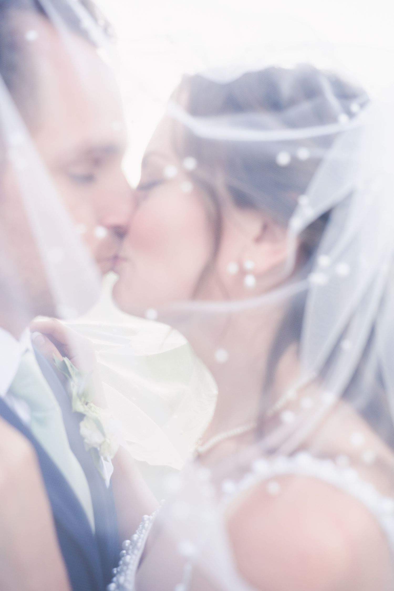 A bride and groom share a blurred kiss under their wedding veil, their faces obscured by the fabric and embedded pearls.