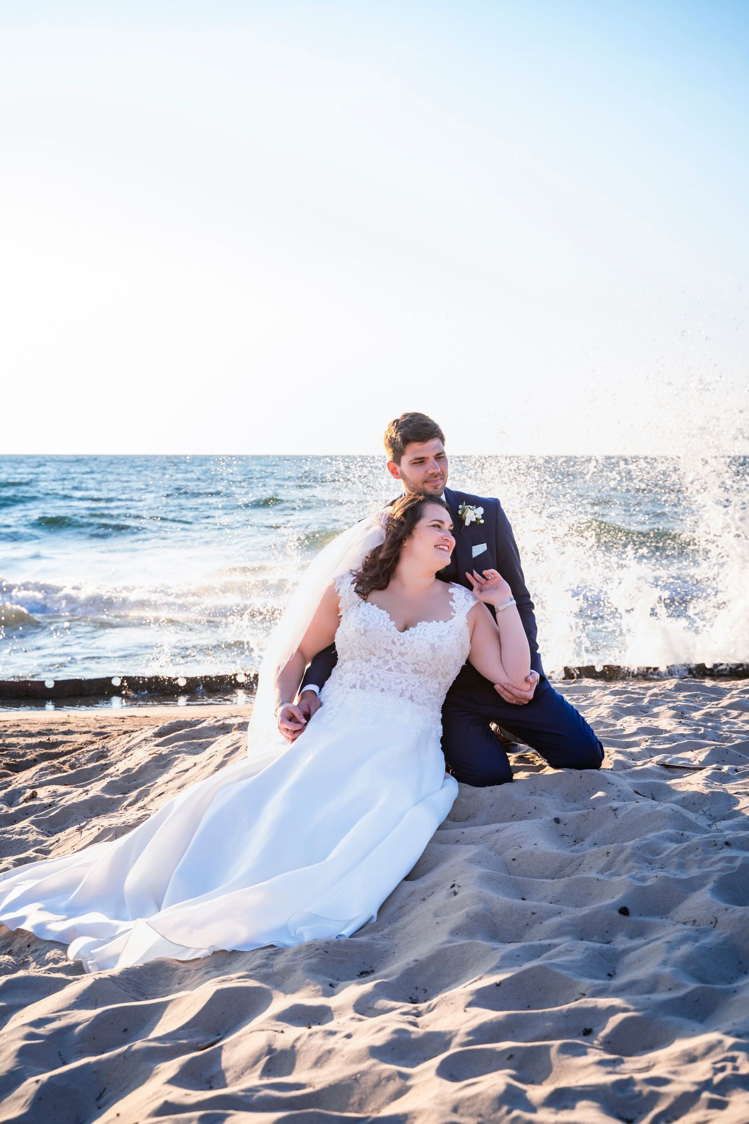 A casual wedding day photograph taken at Lake Michigan of a bride and groom sitting together in the sand on the beach with large waves.