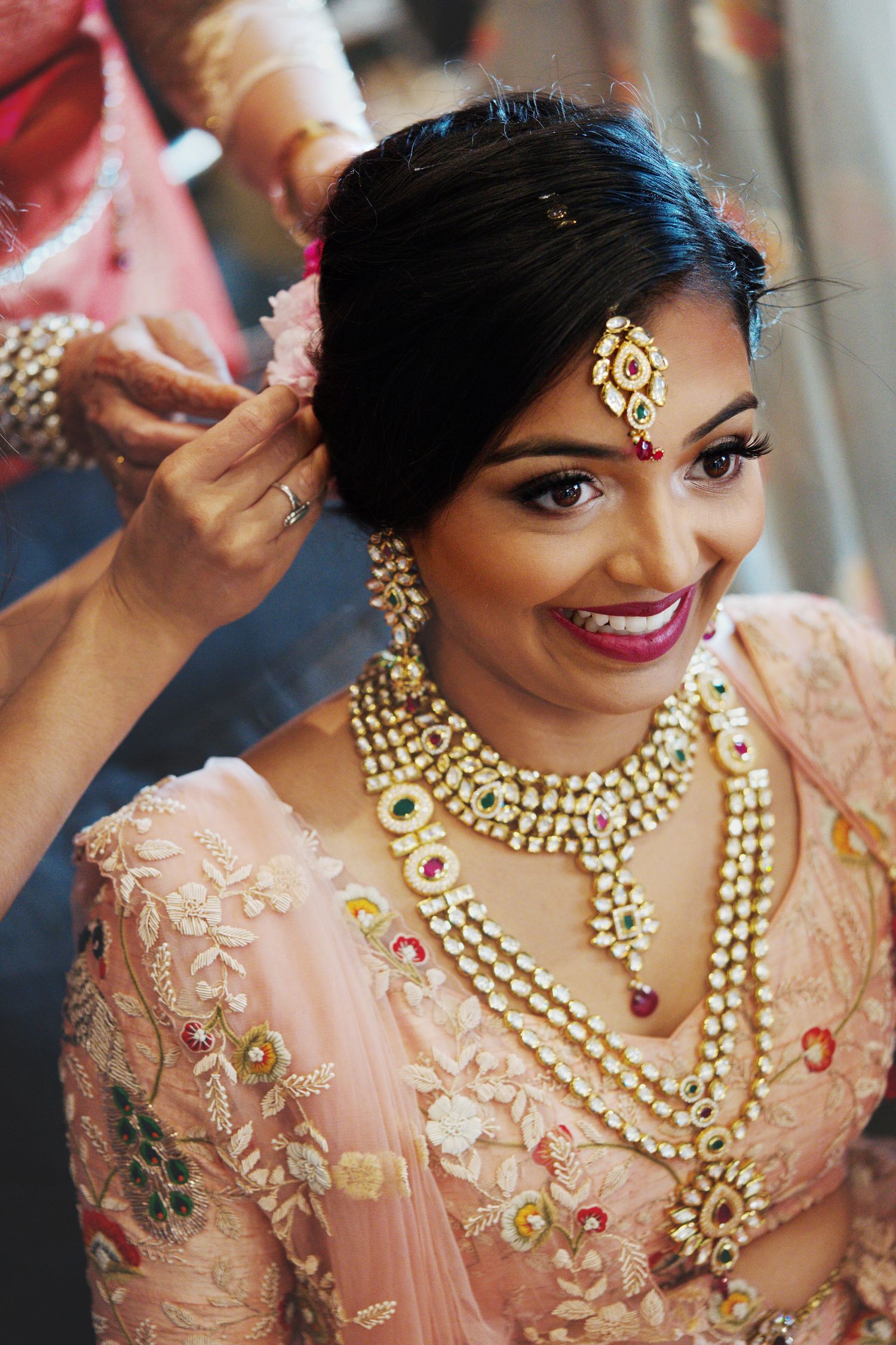 A South East Asian bridal prep scene. The bride smiles before her wedding reception with all her jewelry on, while helpers finish her hair.