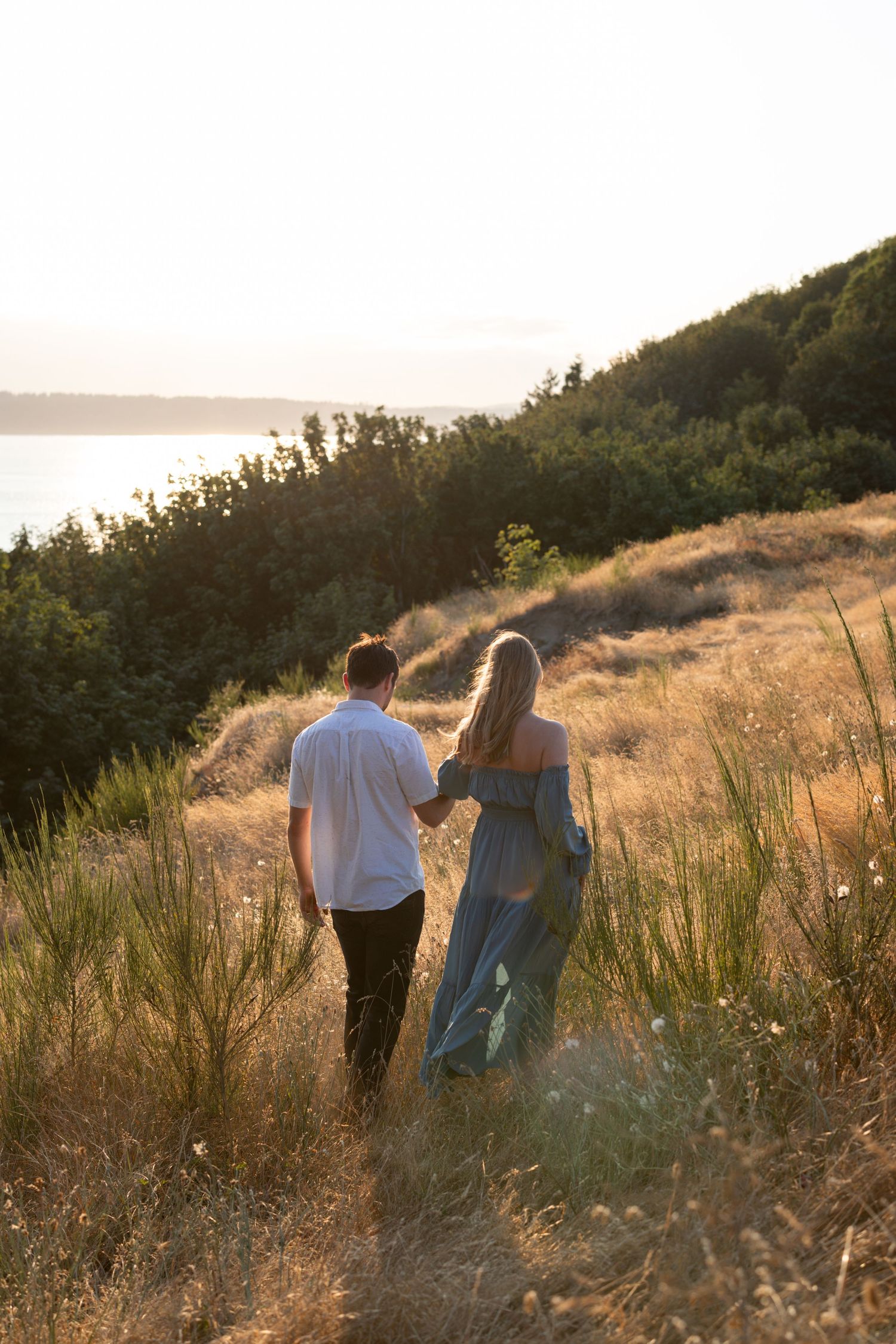 Couple walking along scenic hillside trail during golden hour overlooking water.