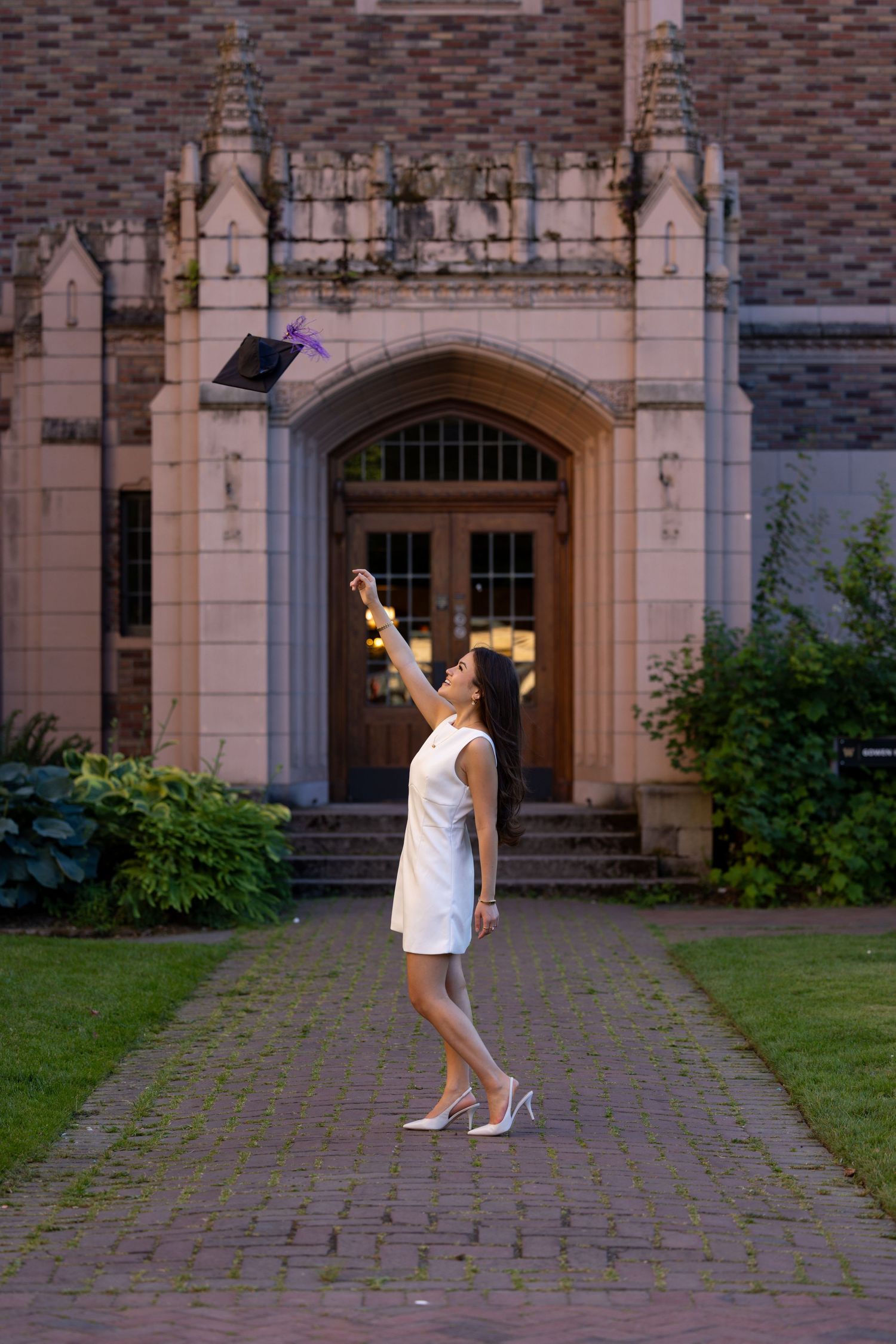 A person in a white dress tossing a graduation cap in front of a Gothic collegiate building entrance.