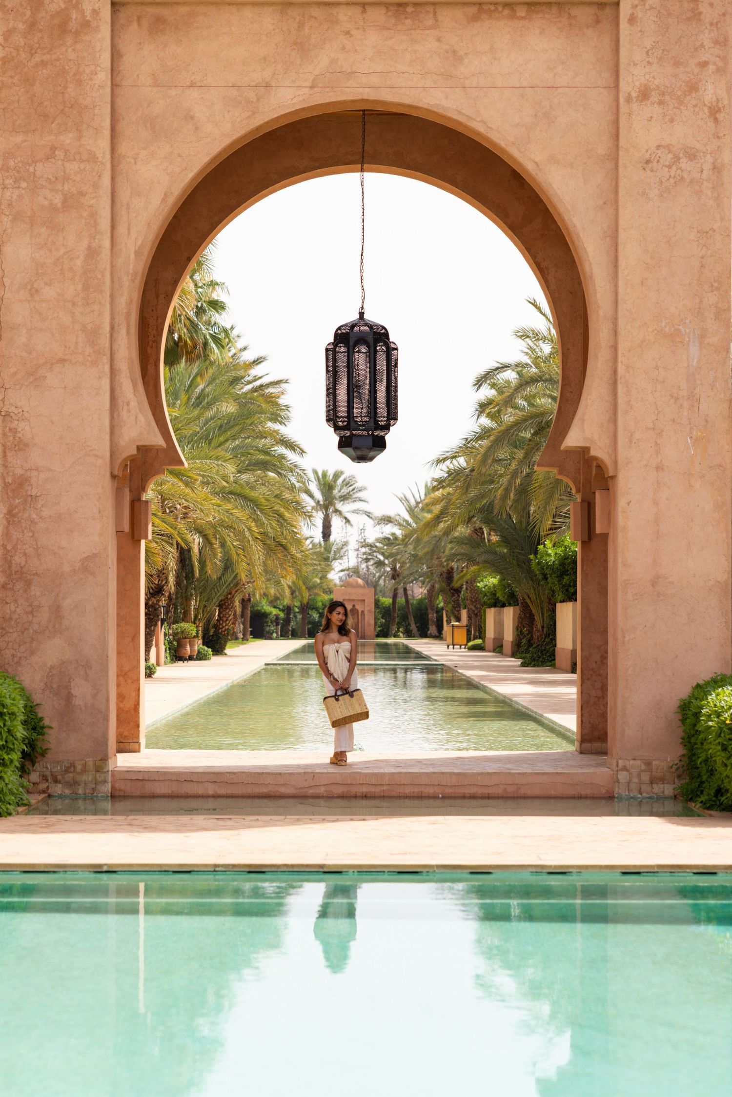 Moroccan architectural archway framing a pool and palm-lined pathway with hanging lantern.
