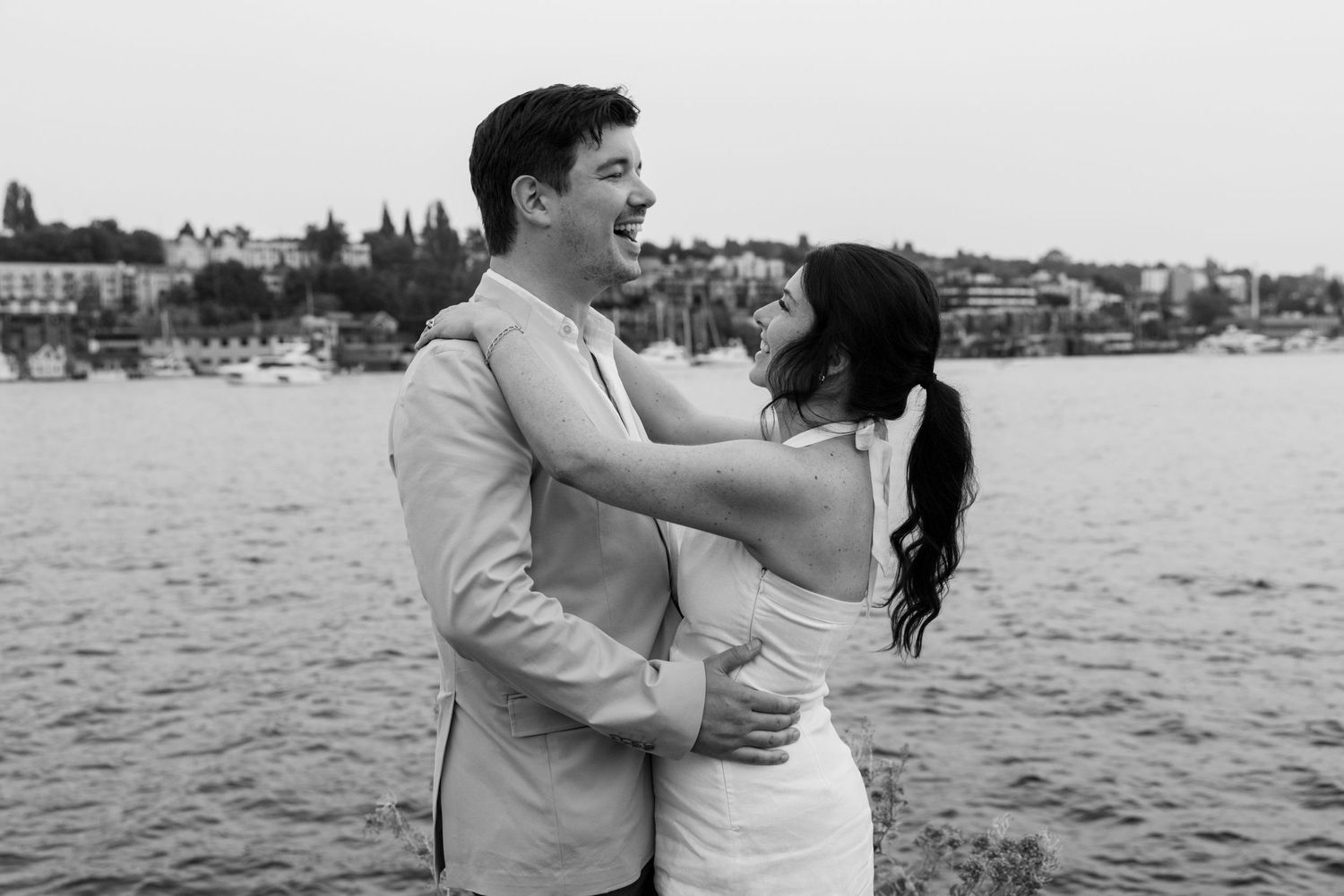 Romantic couple embraces by the waterfront with city skyline in the background.