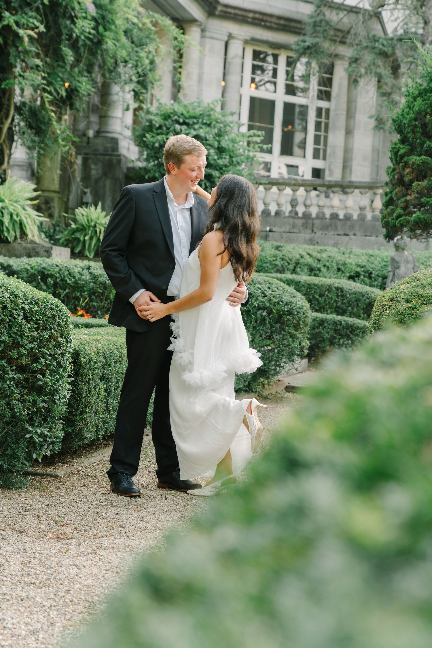 A couple in wedding attire share a joyful moment on a garden path surrounded by manicured hedges and a stone manor.