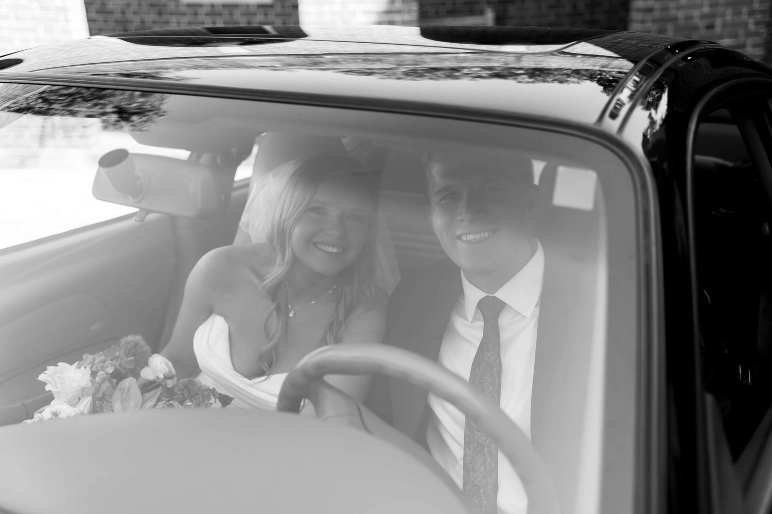 A black and white photo shows a couple smiling while seated in a vintage car during a special occasion.