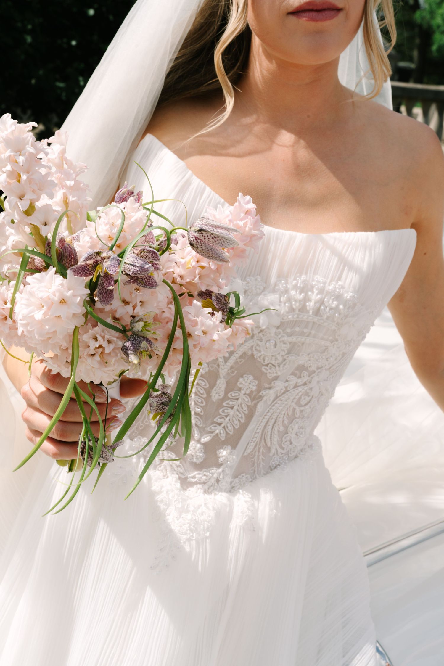 Bride in strapless white lace wedding gown holding elegant pink and purple floral bouquet with green stems outdoors.