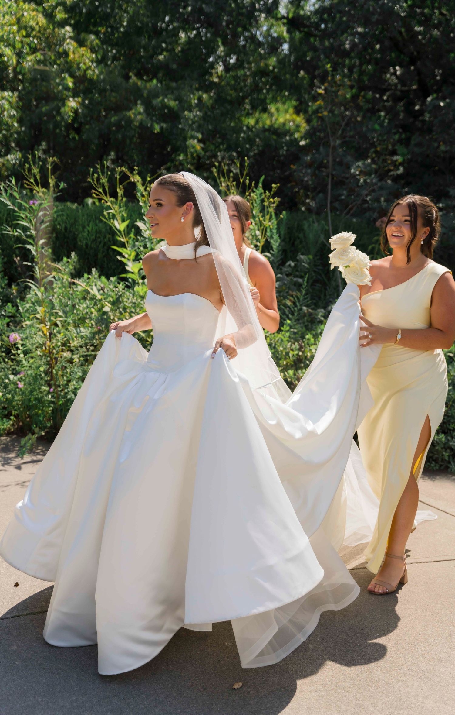 A bride in a flowing white ballgown and veil walks outdoors with a bridesmaid in a yellow dress holding flowers.