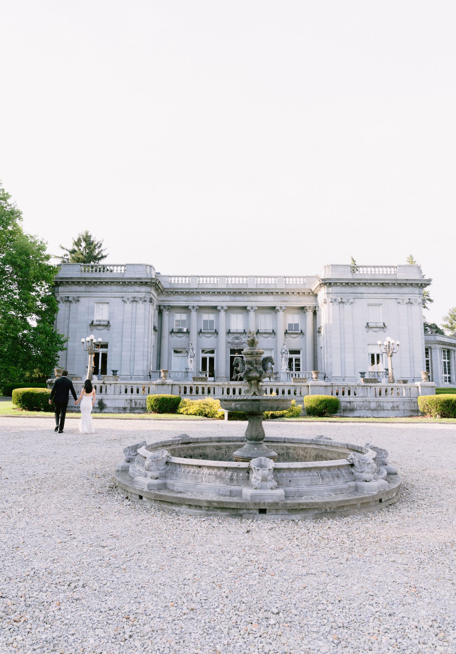 Elegant neoclassical mansion with stone fountain in gravel courtyard, surrounded by lush trees at a luxury wedding venue.