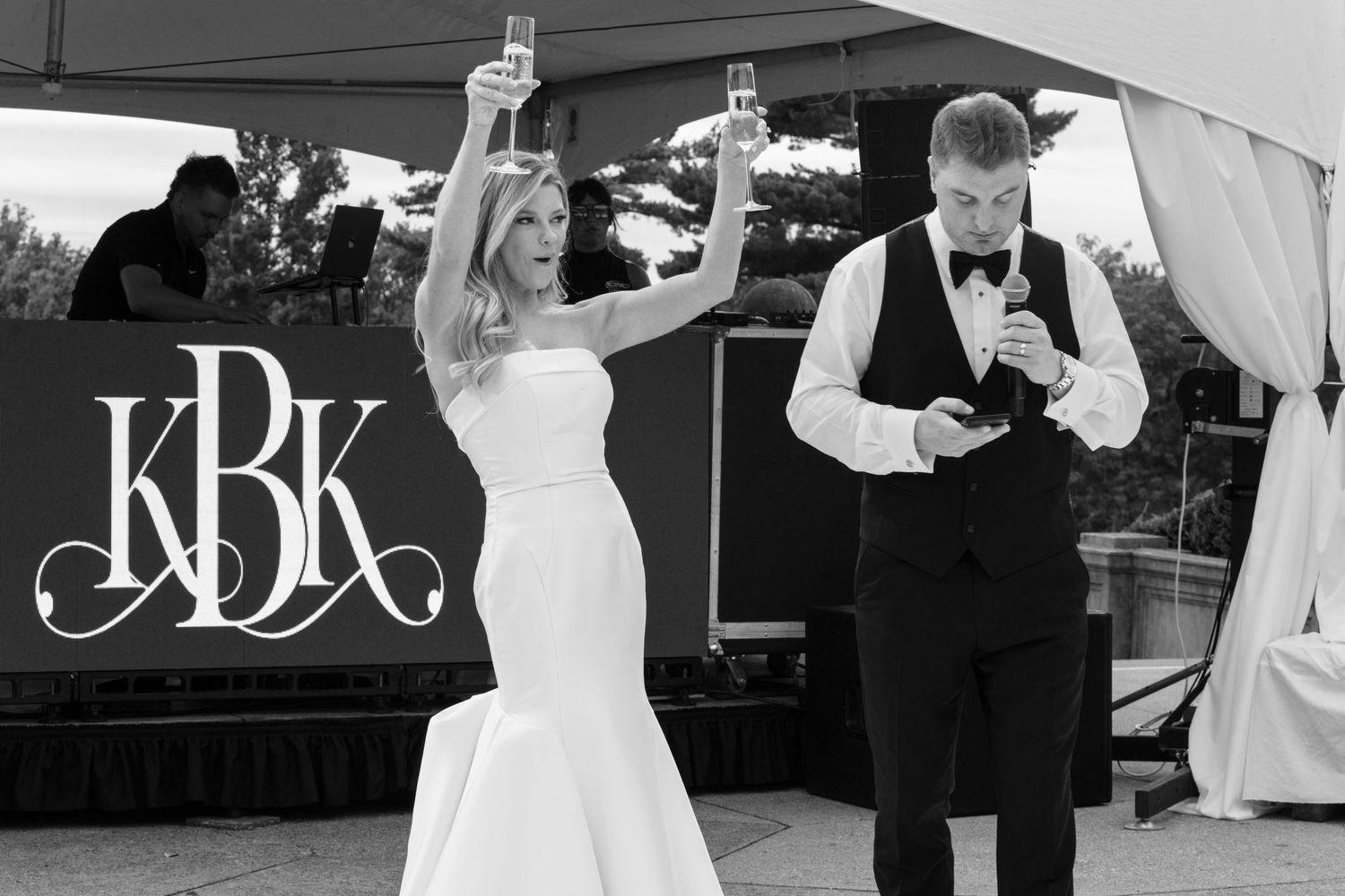 Bride and groom toasting with champagne at outdoor wedding reception with monogram KBK backdrop.