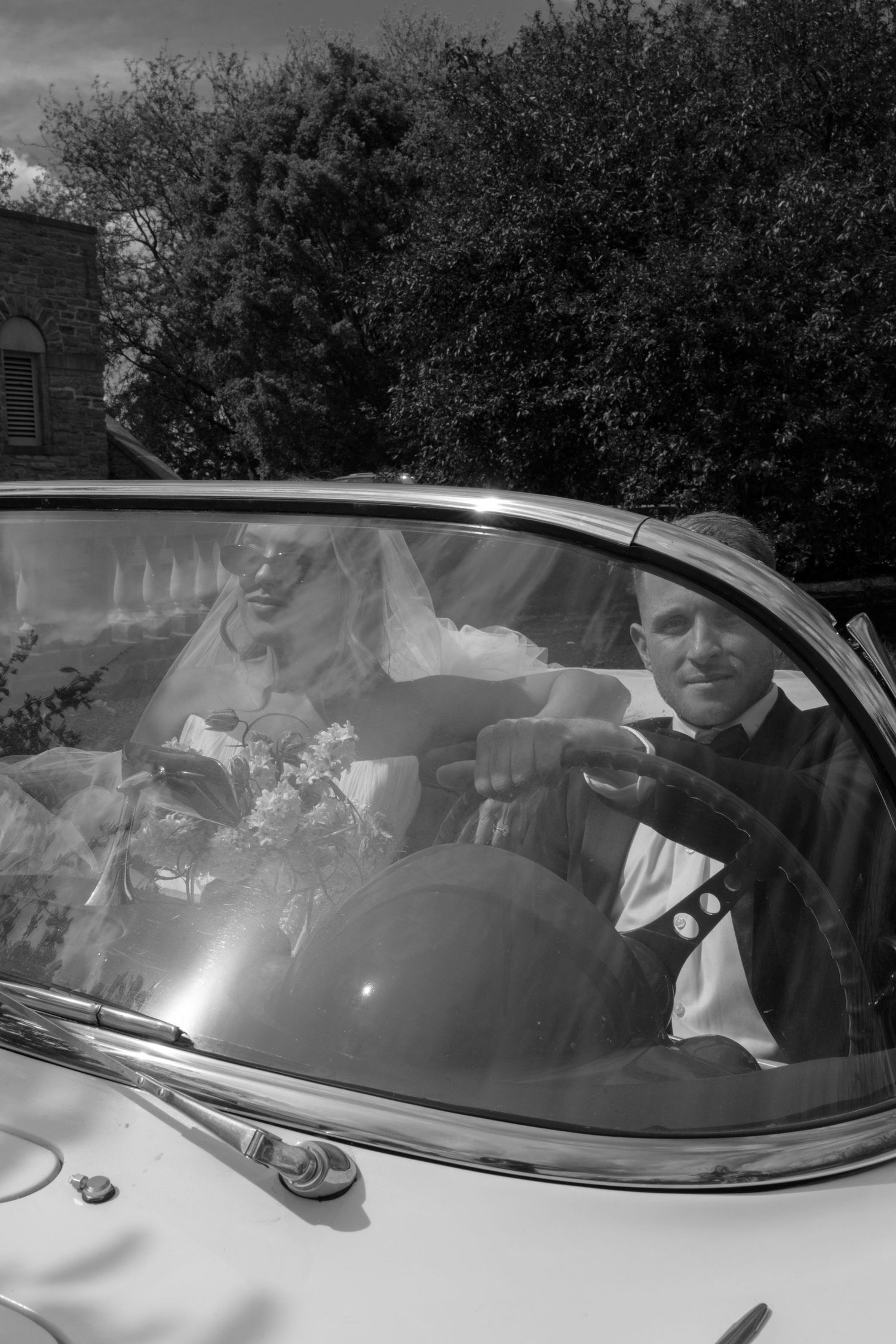 Black and white photo of a bride and groom sitting in a vintage convertible car on their wedding day.