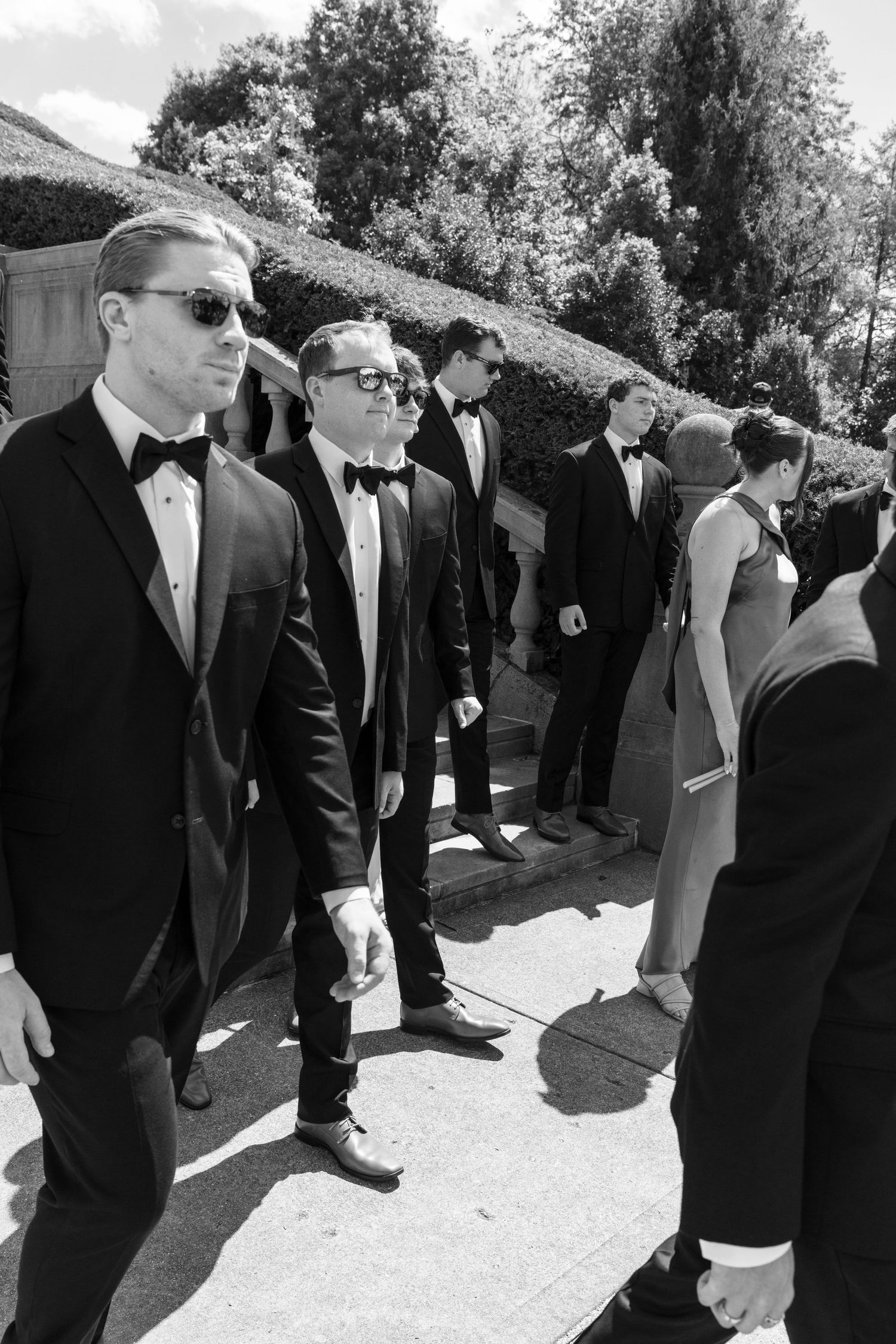 Black and white photo of groomsmen in tuxedos and bow ties gathered outdoors at a formal wedding event.