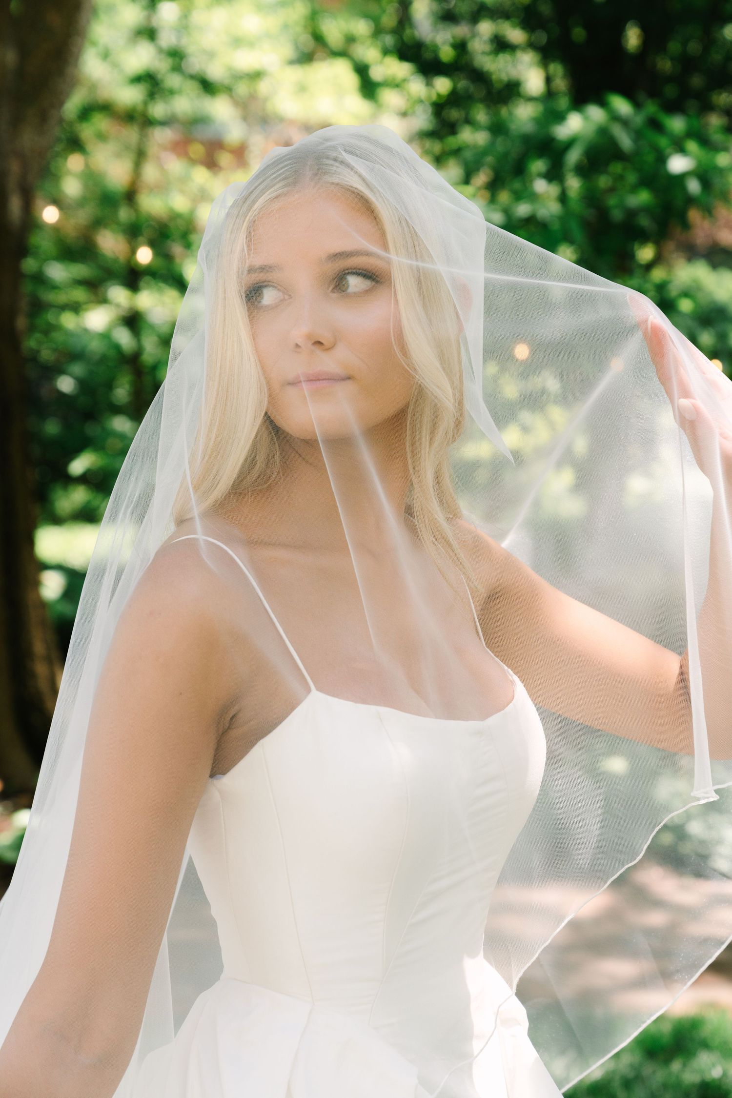 Blonde bride in white gown lifts sheer veil outdoors, surrounded by lush green foliage in soft natural light.