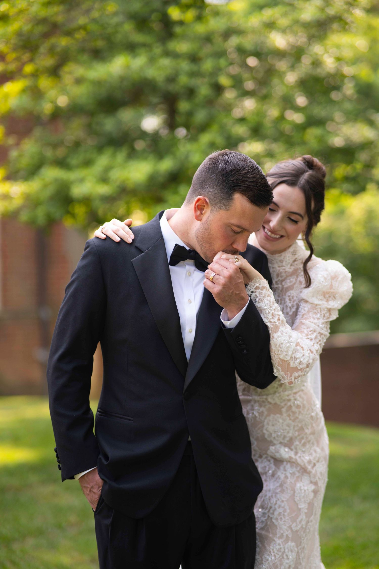 A romantic wedding moment in an outdoor garden setting, with a black tuxedo and lace dress against natural greenery.