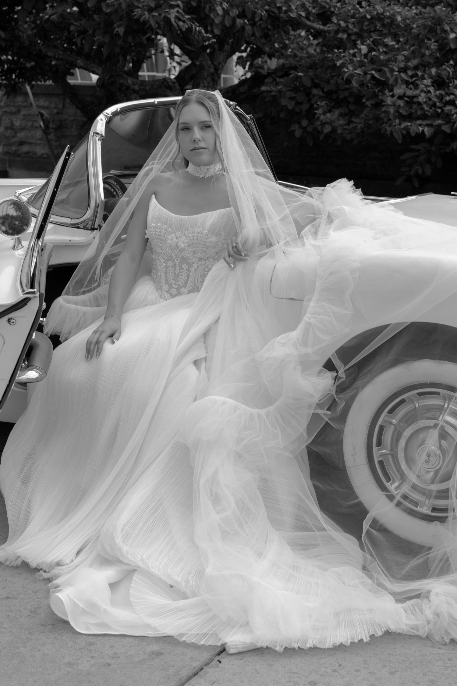 Black and white photo of a bride in a flowing wedding gown and veil posed beside a vintage classic car.