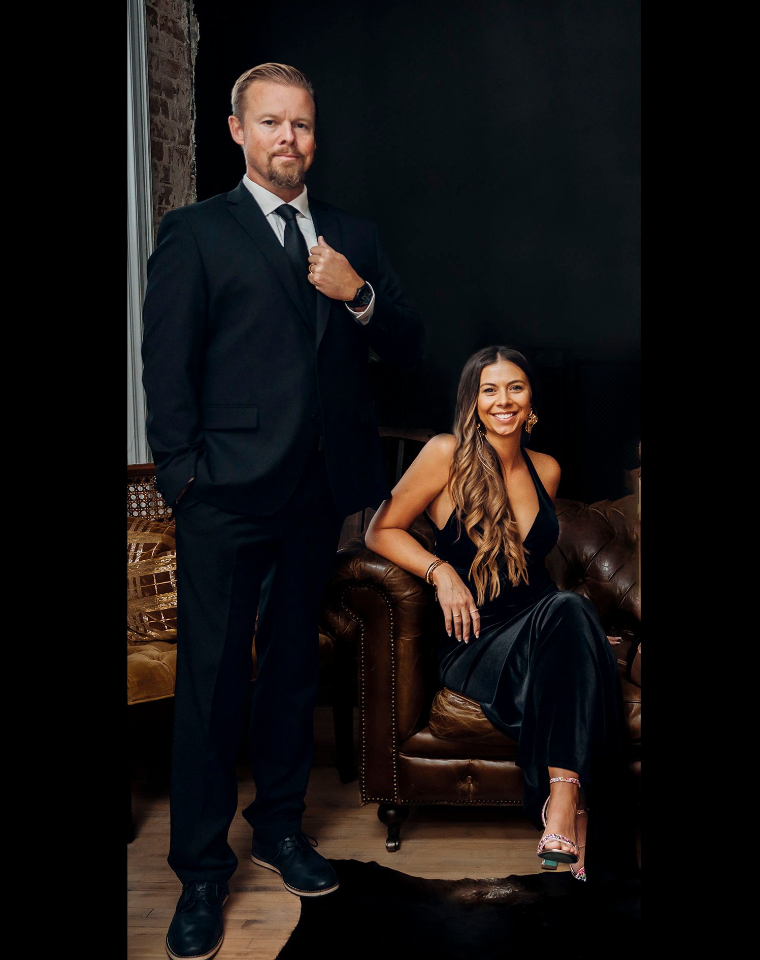 Professional studio portrait of a couple in formal black attire posing together against a dark background.