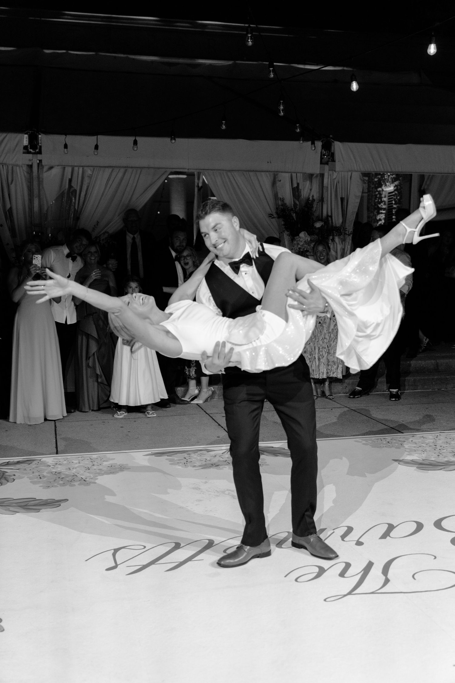 Groom lifts bride during first dance at elegant wedding reception in black and white photo.
