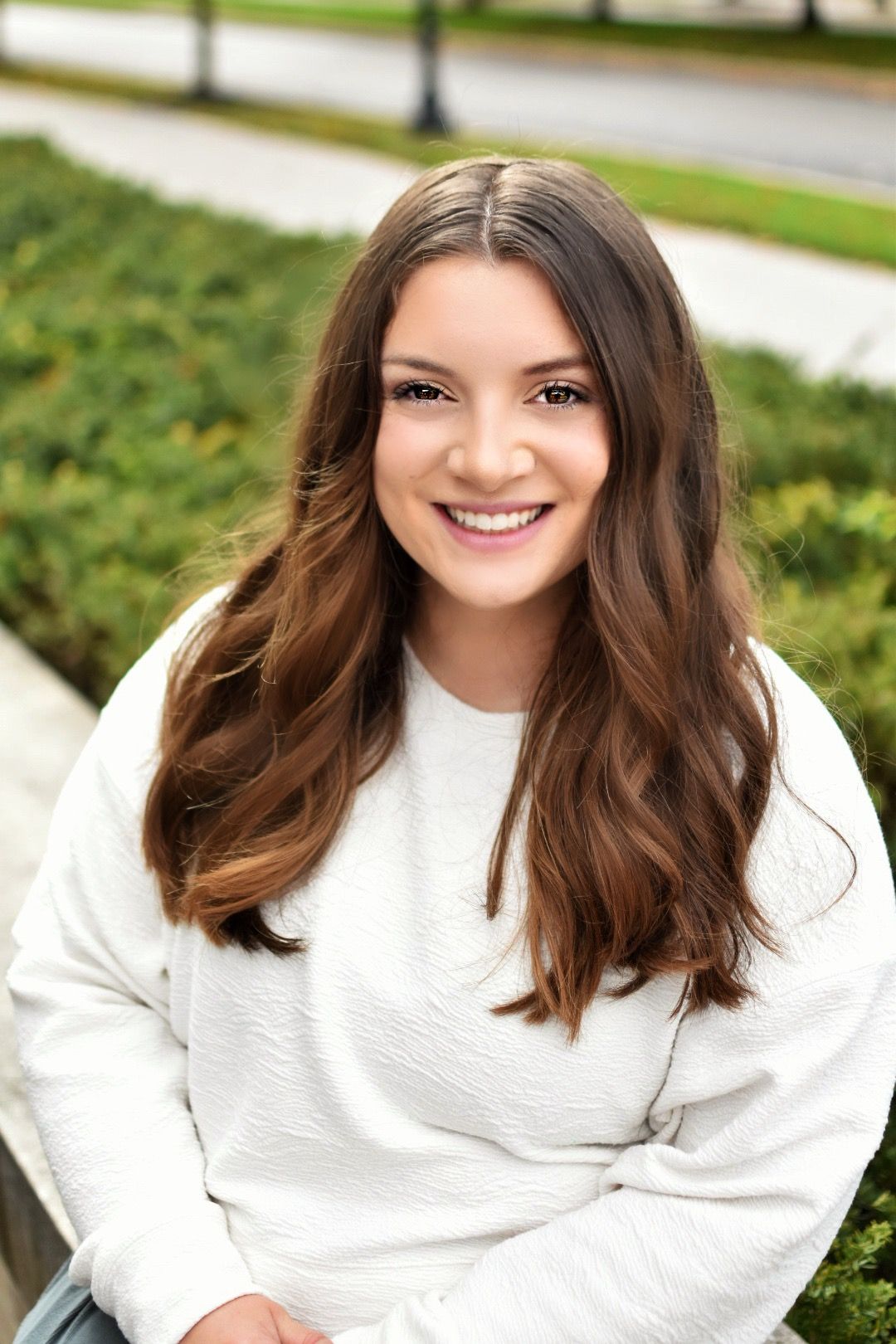 Smiling young woman with long brown hair wearing a white sweater, sitting outdoors near green hedges.