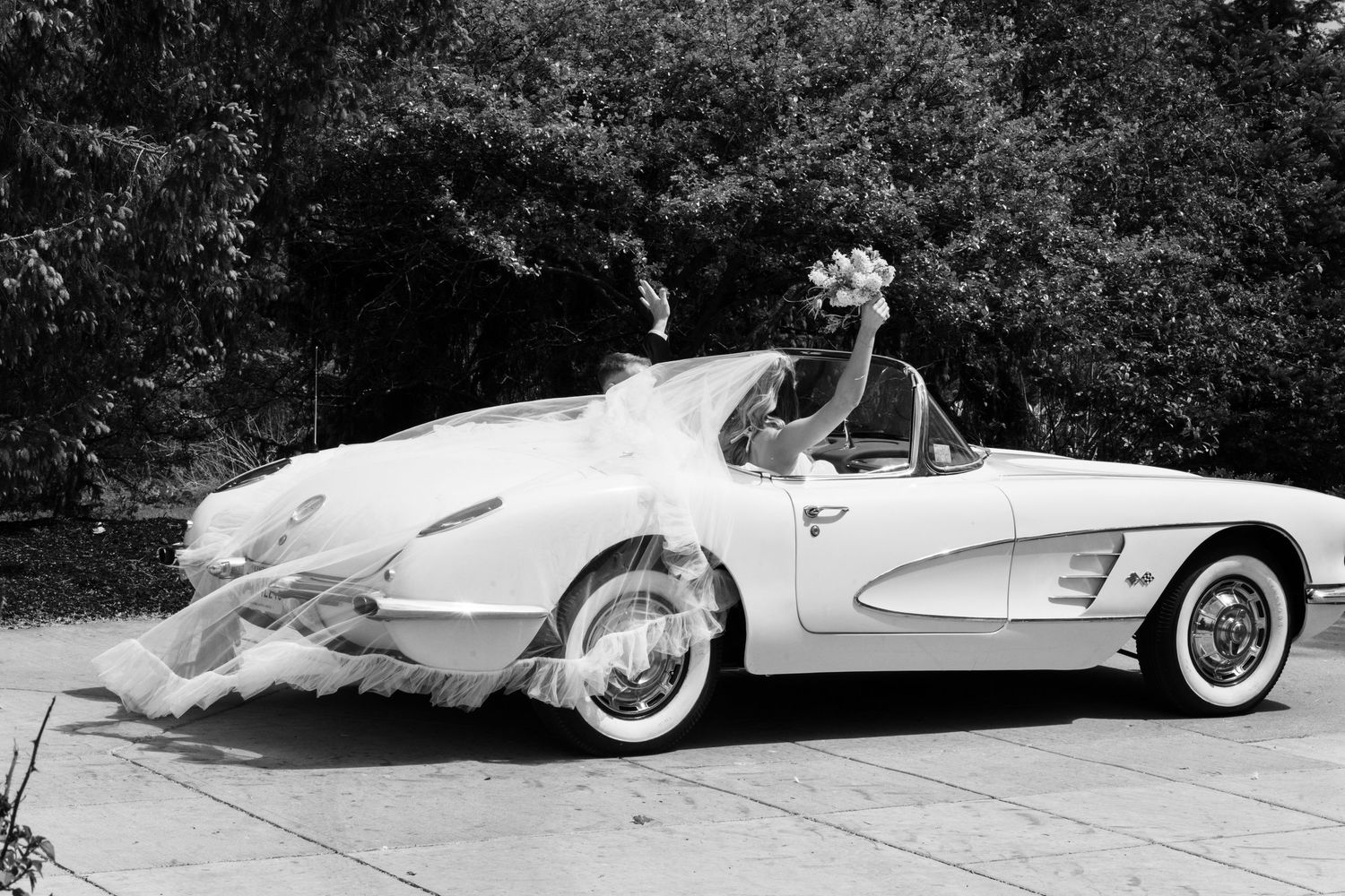 Bride in wedding dress riding in a classic white 1959 Chevrolet Corvette convertible, black and white photo.