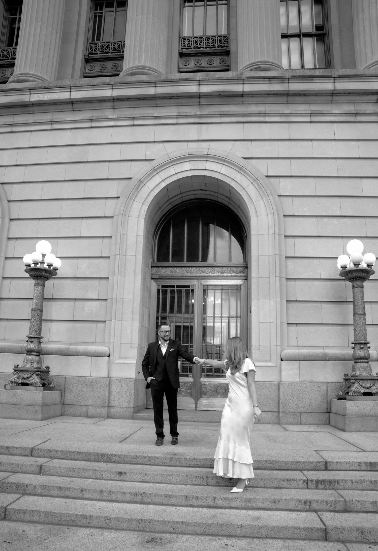 Black and white photo of a couple on steps of a grand neoclassical building with ornate lamp posts.