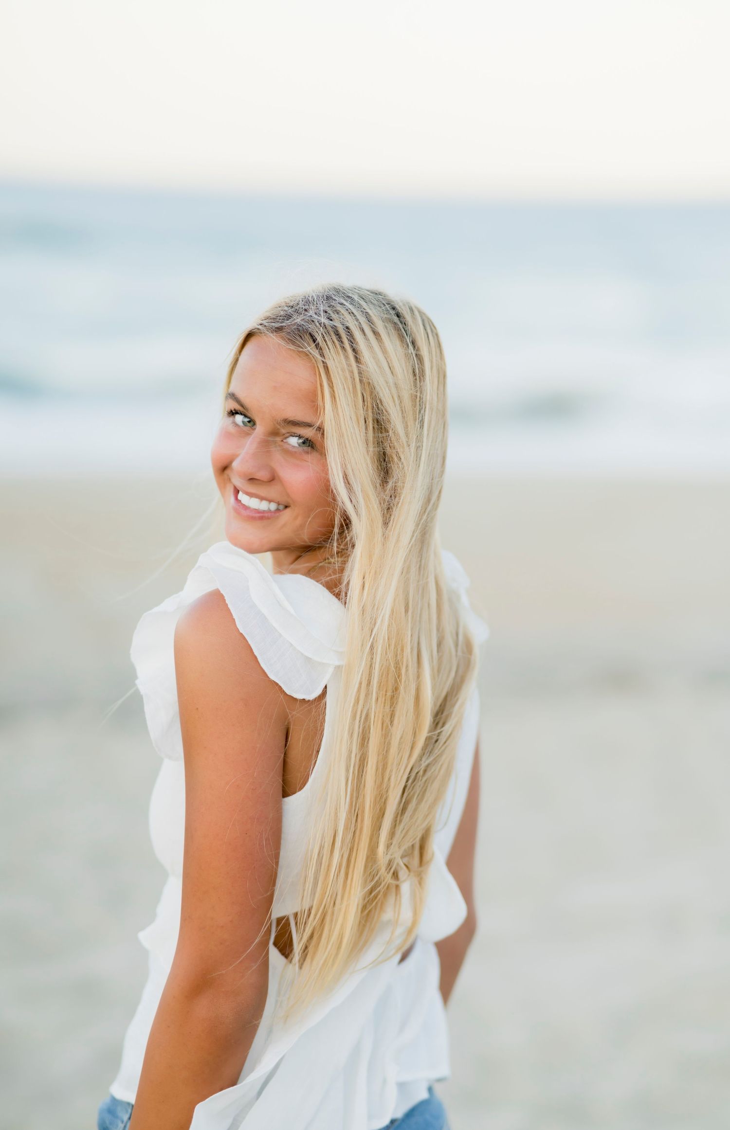 A person in a white ruffled top and blonde hair poses at the beach with a joyful expression.
