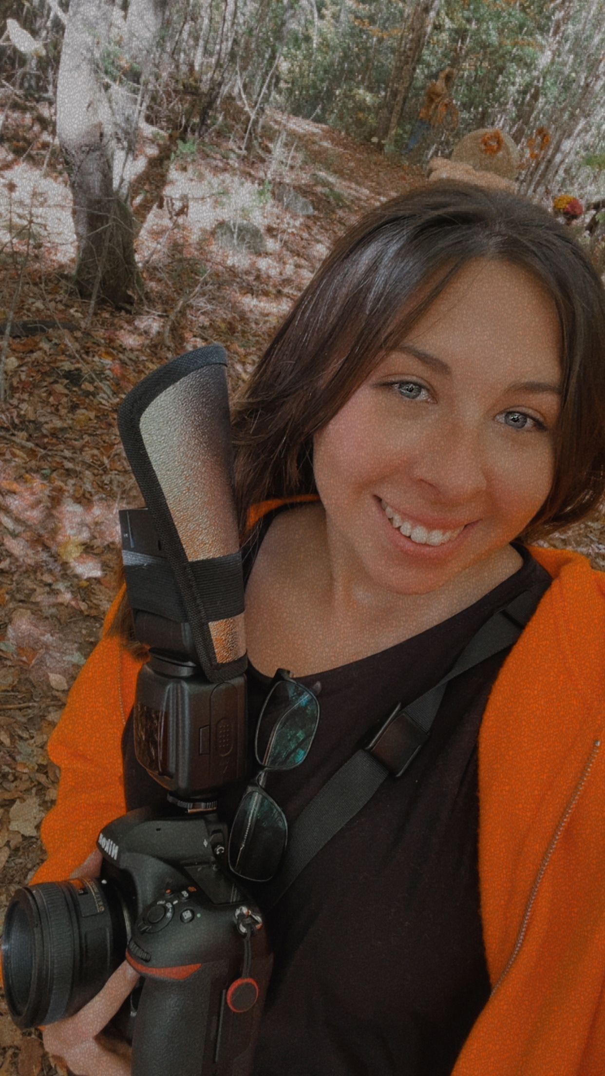 A photographer with a professional camera and flash setup smiles while taking selfies against a brick wall background.