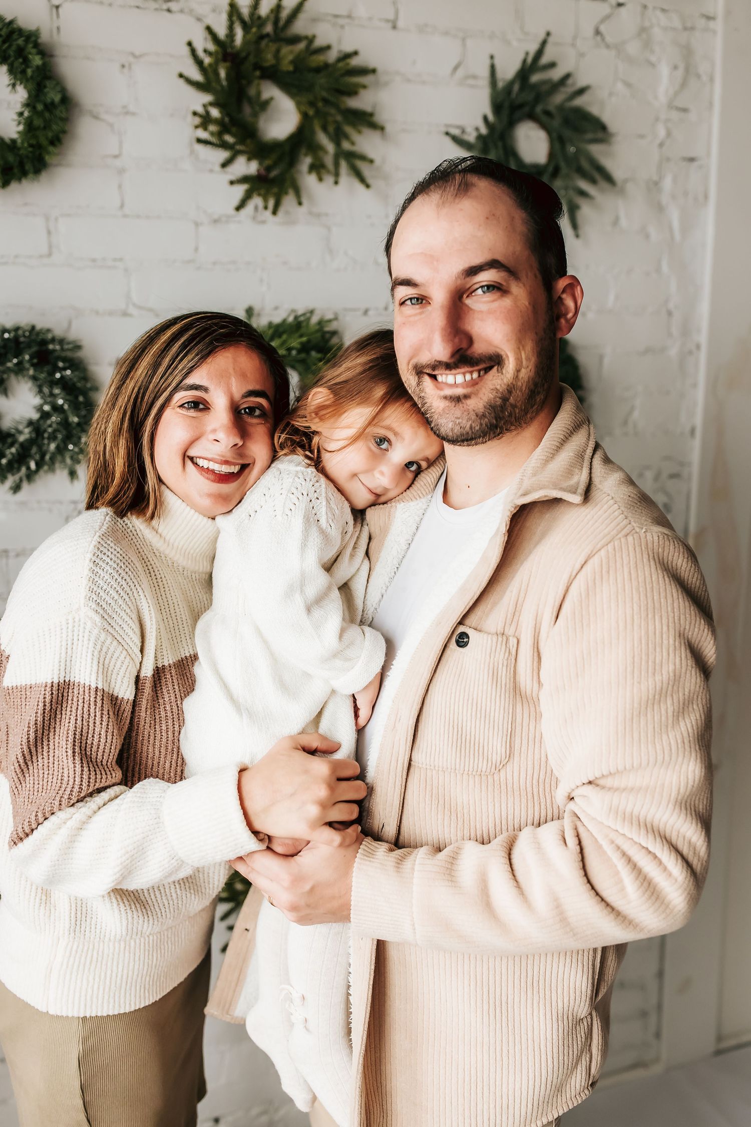 A family wearing cozy neutral sweaters pose together against a white brick wall with holiday wreaths in the background.