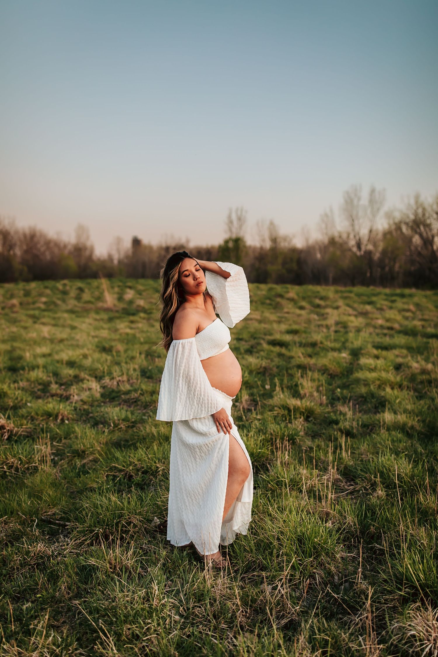 A, expecting woman in a flowing white dress poses in a sunlit meadow during golden hour with trees in the background.