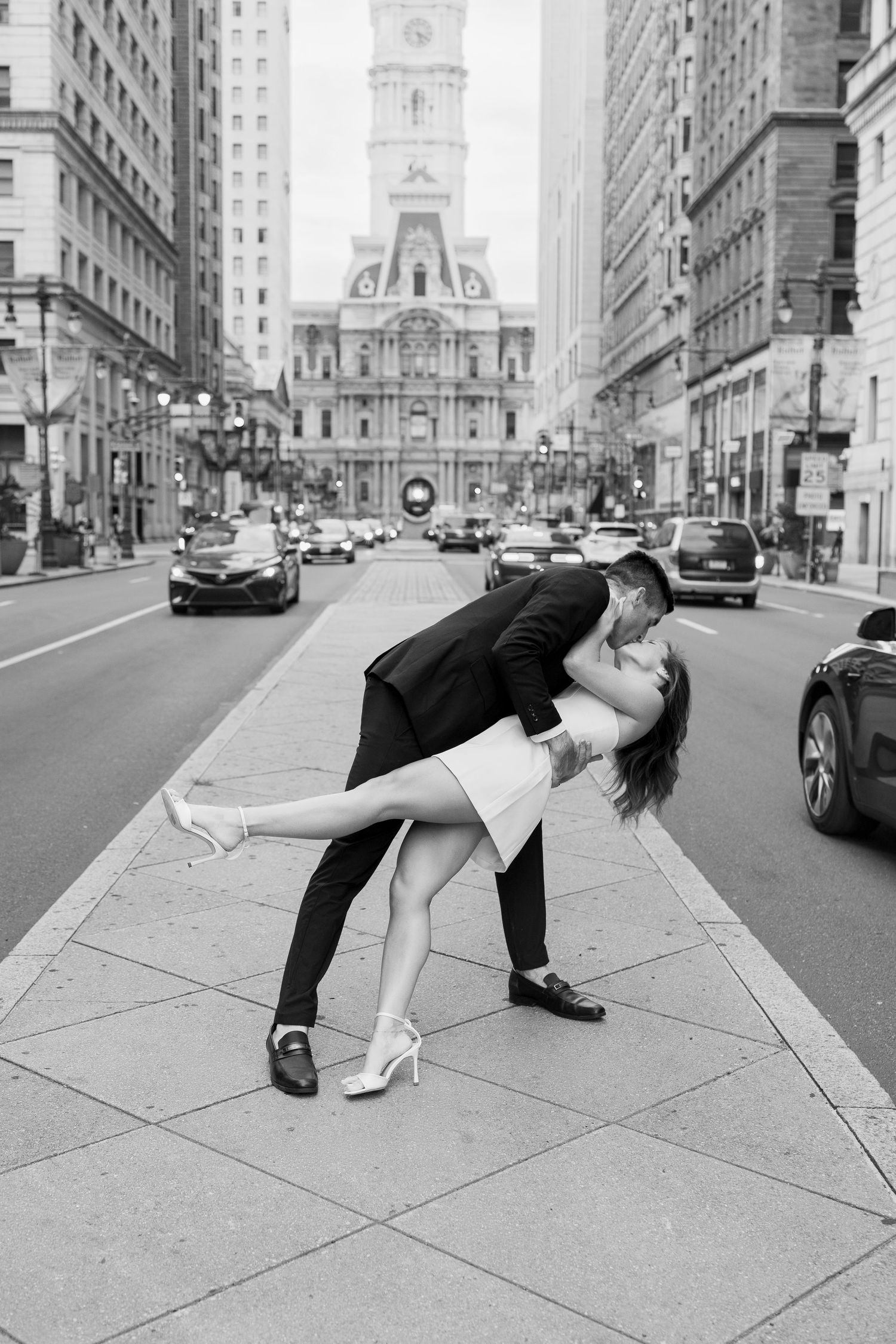 Black-and-white engagement photo of a couple kissing on a Philadelphia sidewalk with downtown architecture in the background.