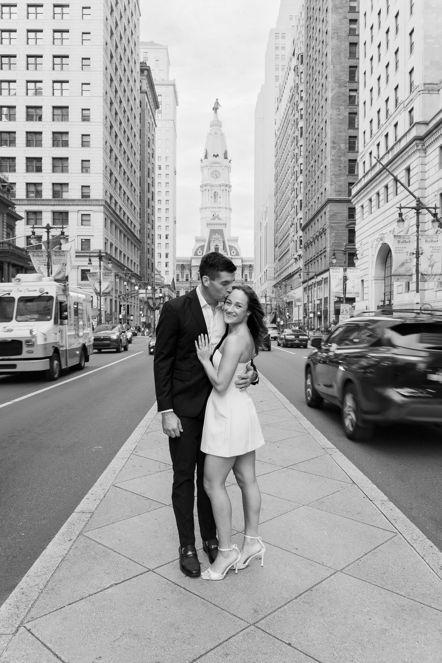 Black-and-white engagement photo of a couple kissing on a Philadelphia sidewalk with downtown architecture in the background.