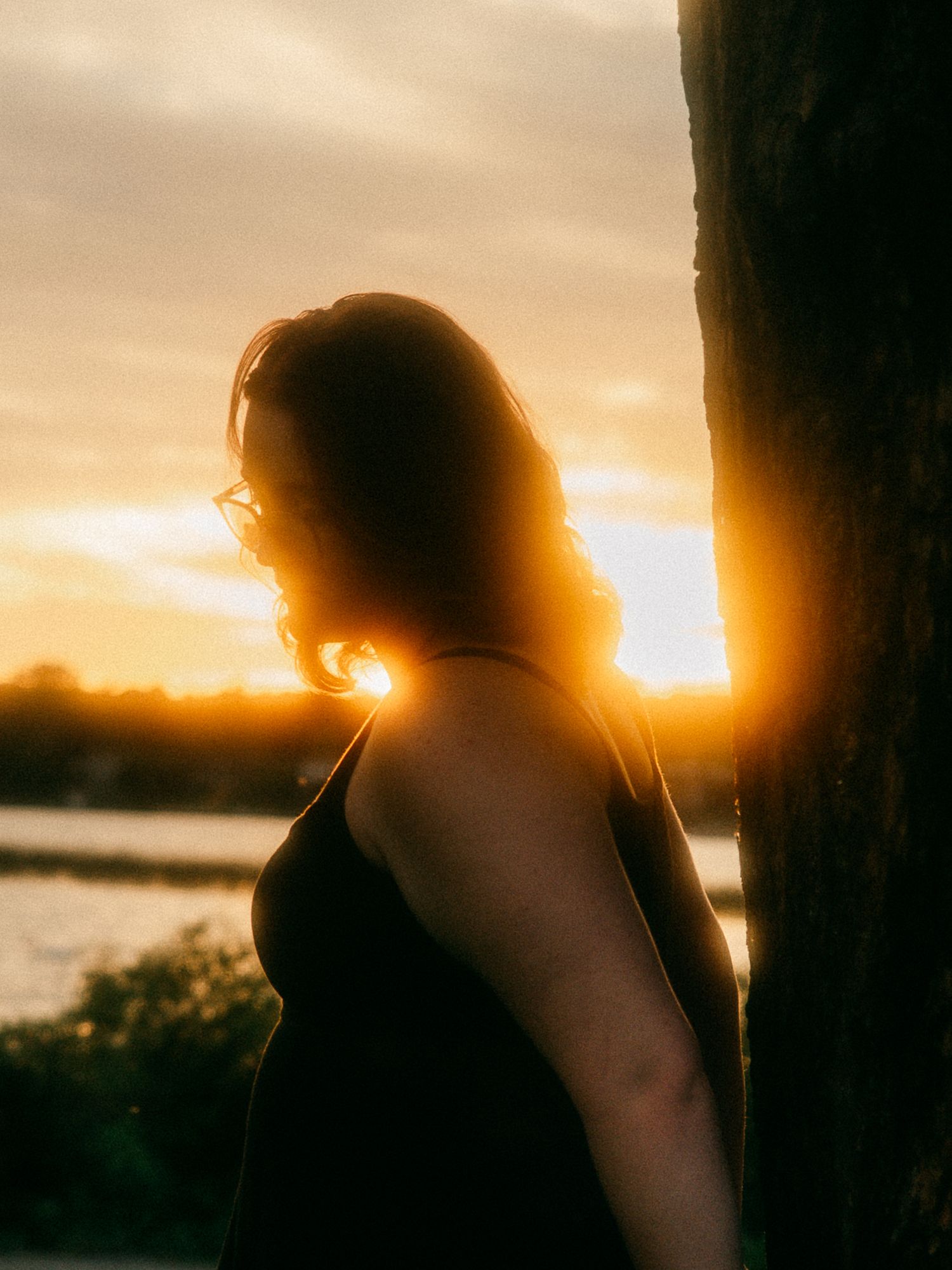 Silhouette of woman beside tree with golden sunset glowing behind her near a lake.