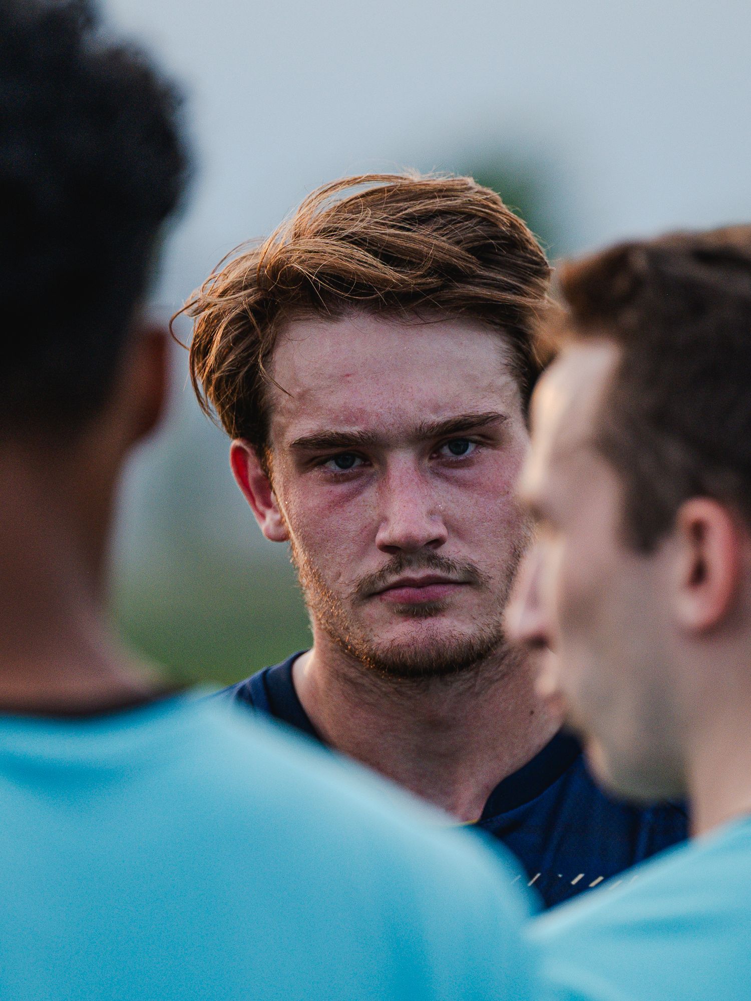 Close-up of a focused red-haired soccer player in a light blue jersey during a team huddle.