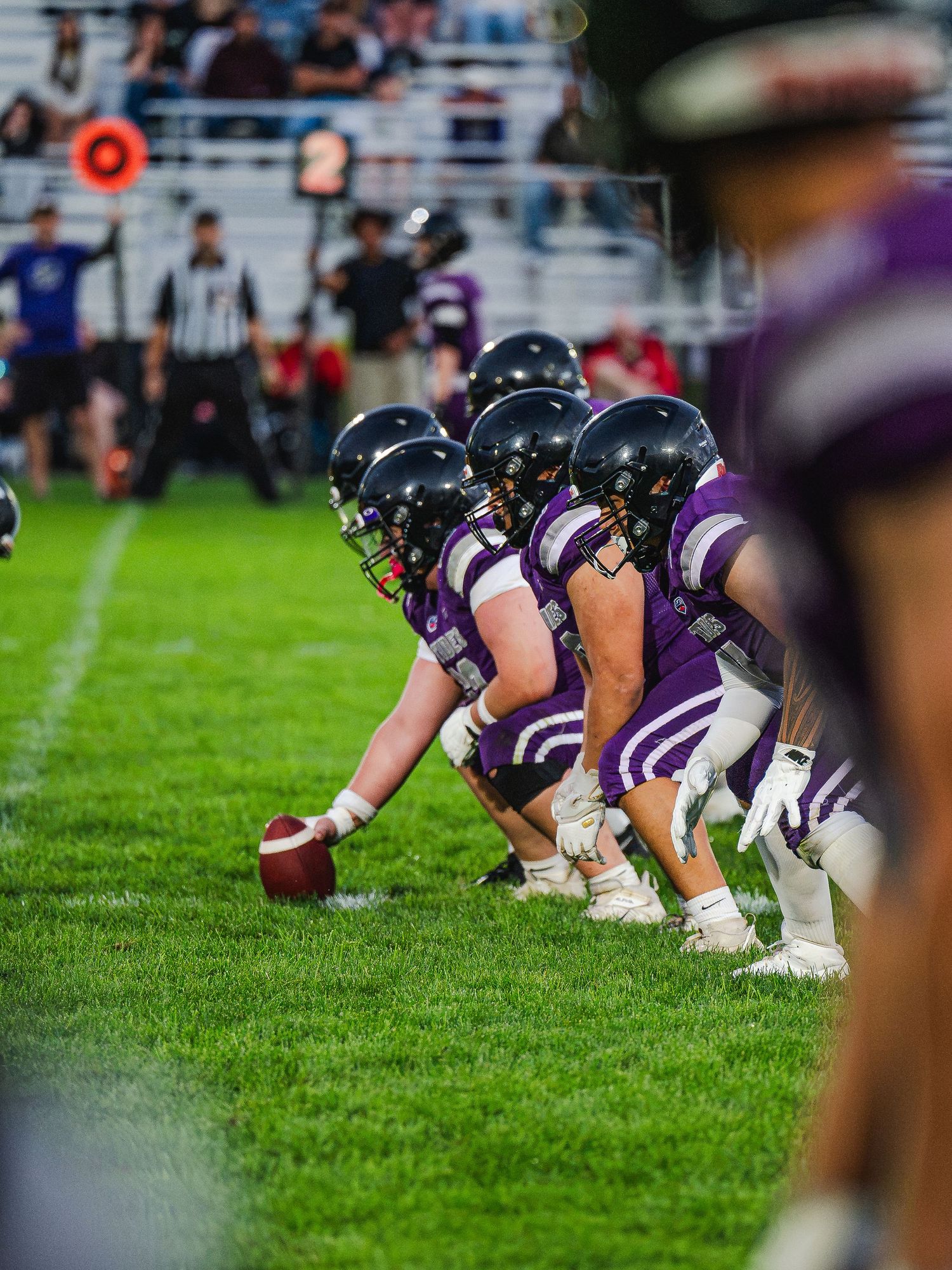 Riptides football offensive line prepares for snap in purple uniforms under stadium lights.