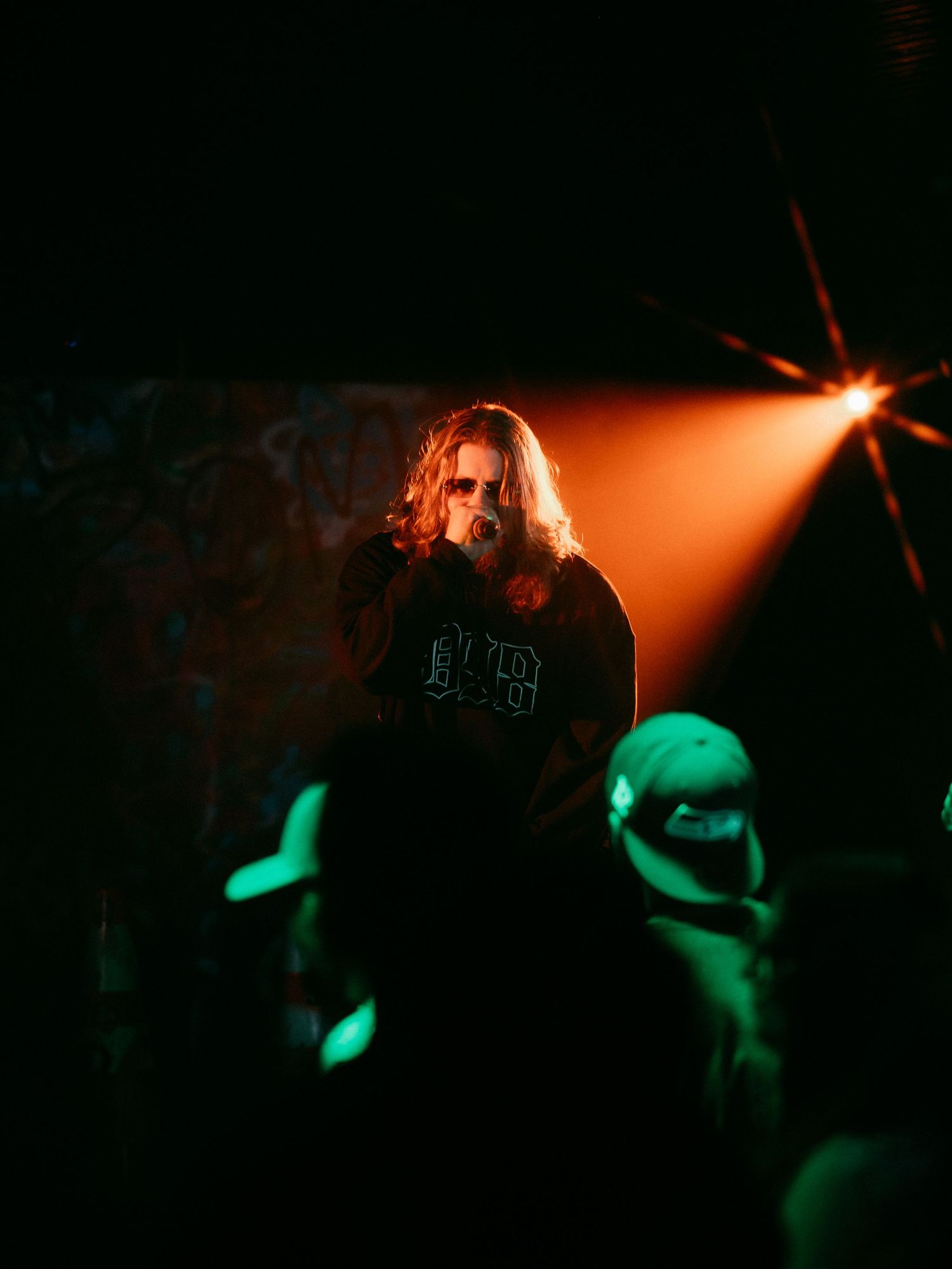 Long-haired performer singing on stage under dramatic orange spotlight in dark concert venue.
