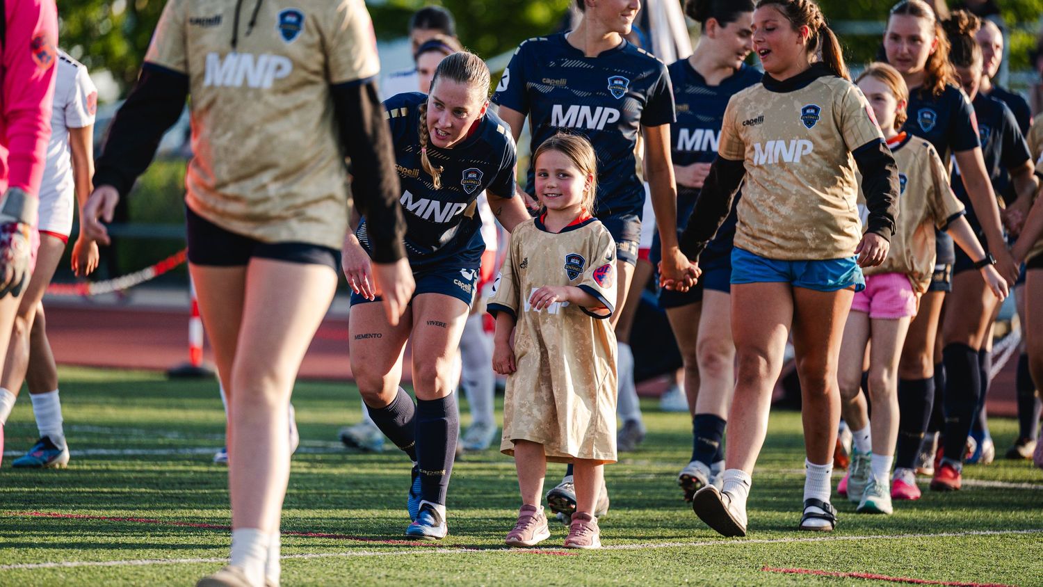 Young child walks hand-in-hand with female soccer players in dark uniforms during a pre-game ceremony on the field.