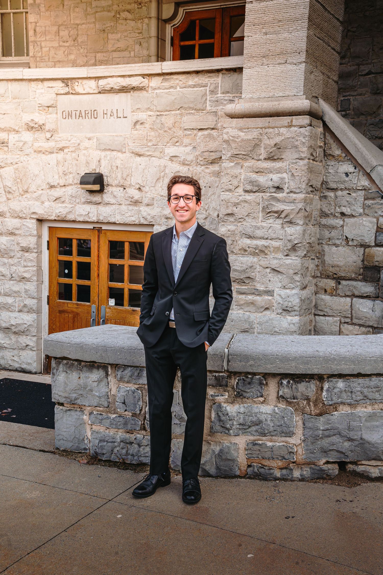 Young man in black suit stands confidently outside Ontario Hall stone university building.