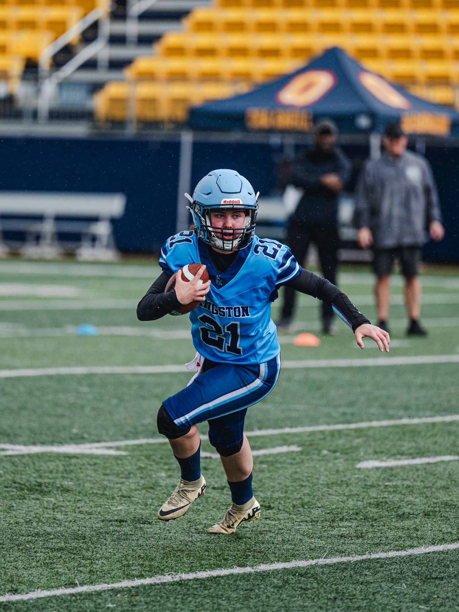 Young football player number 21 in a light blue Kingston uniform runs with the ball on a stadium field.