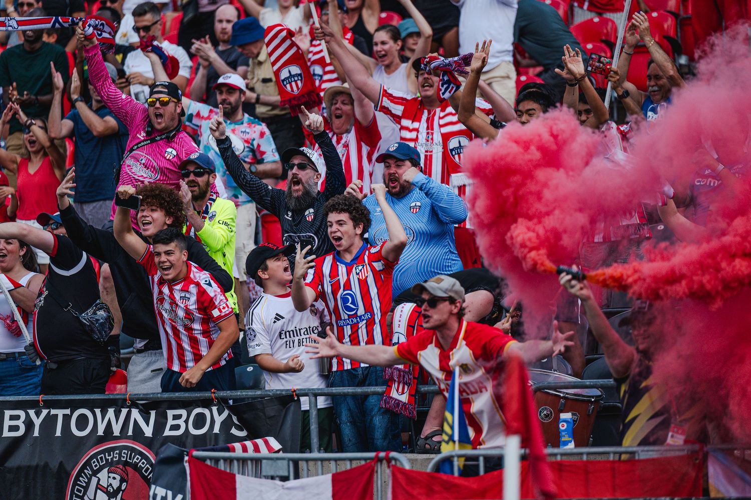 Passionate soccer fans celebrating with red smoke flare at a match near Bytown Boys banner.