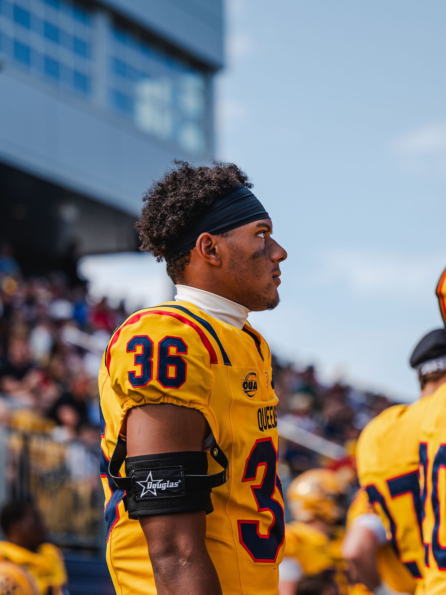 Queen's football player #36 in gold uniform with headband stands on sideline during college football game.