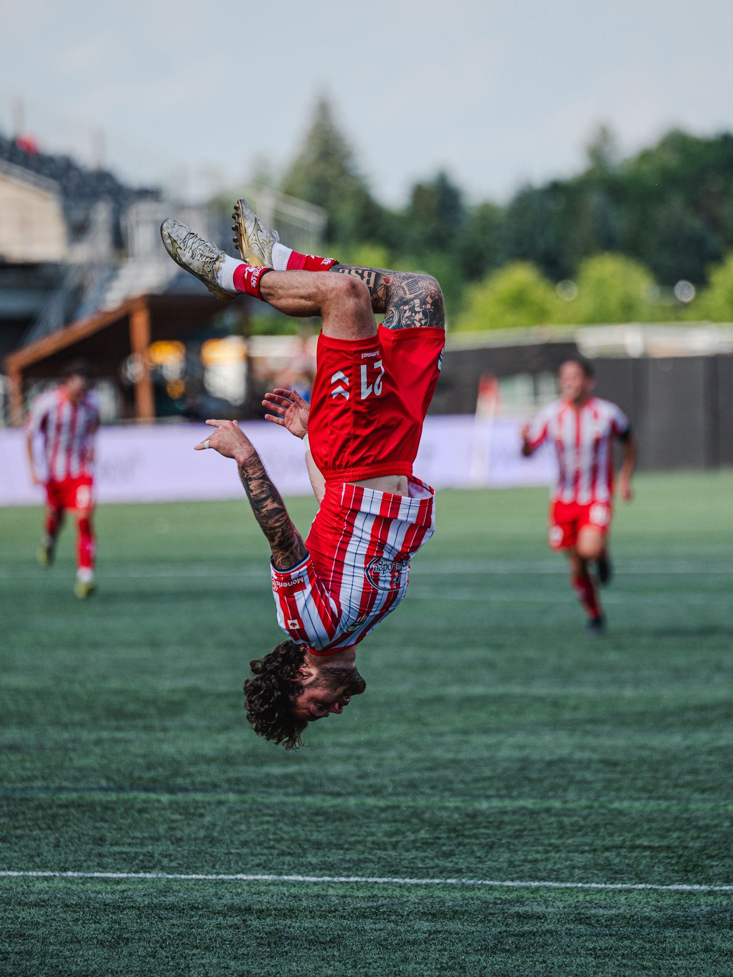 A tattooed soccer player in a red uniform performs a dramatic backflip celebration on a green field during a match.