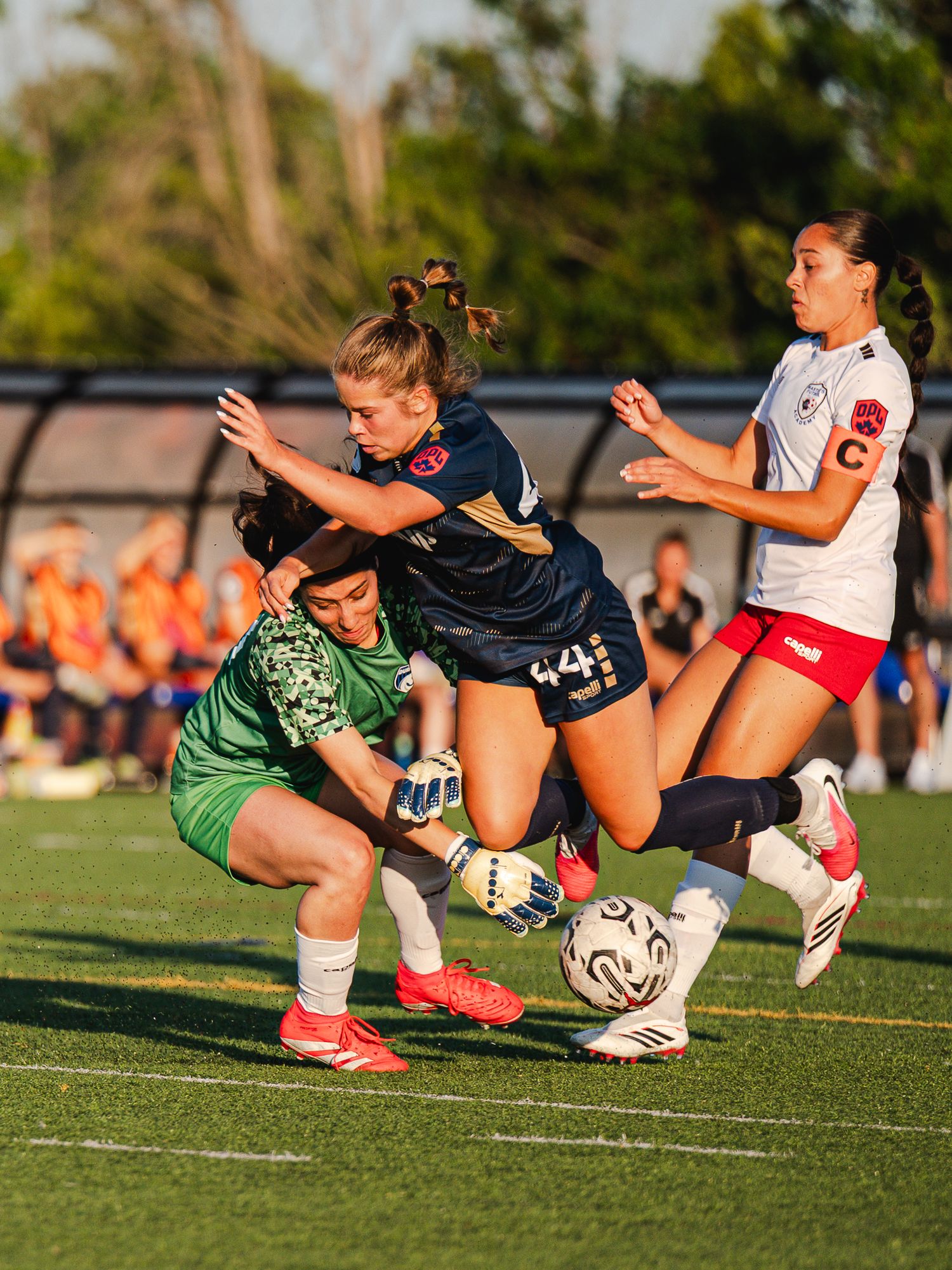 Female soccer players compete for the ball as a goalkeeper in green attempts to make a save during a match.