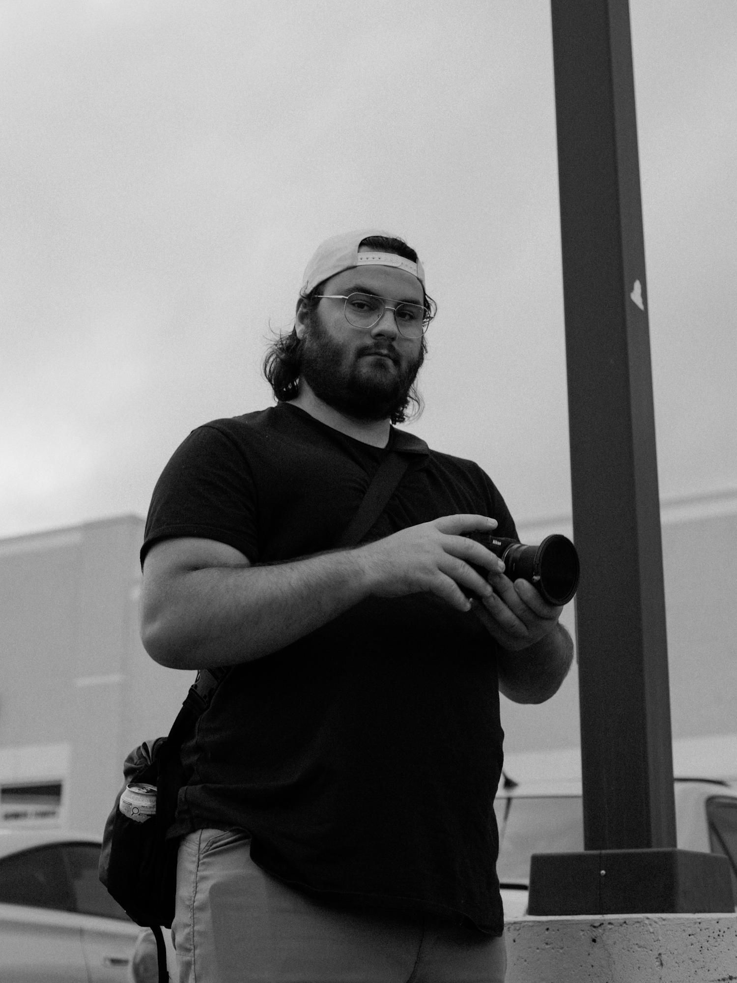 Black and white photo of a bearded man wearing glasses and a backwards cap, holding a camera outdoors.